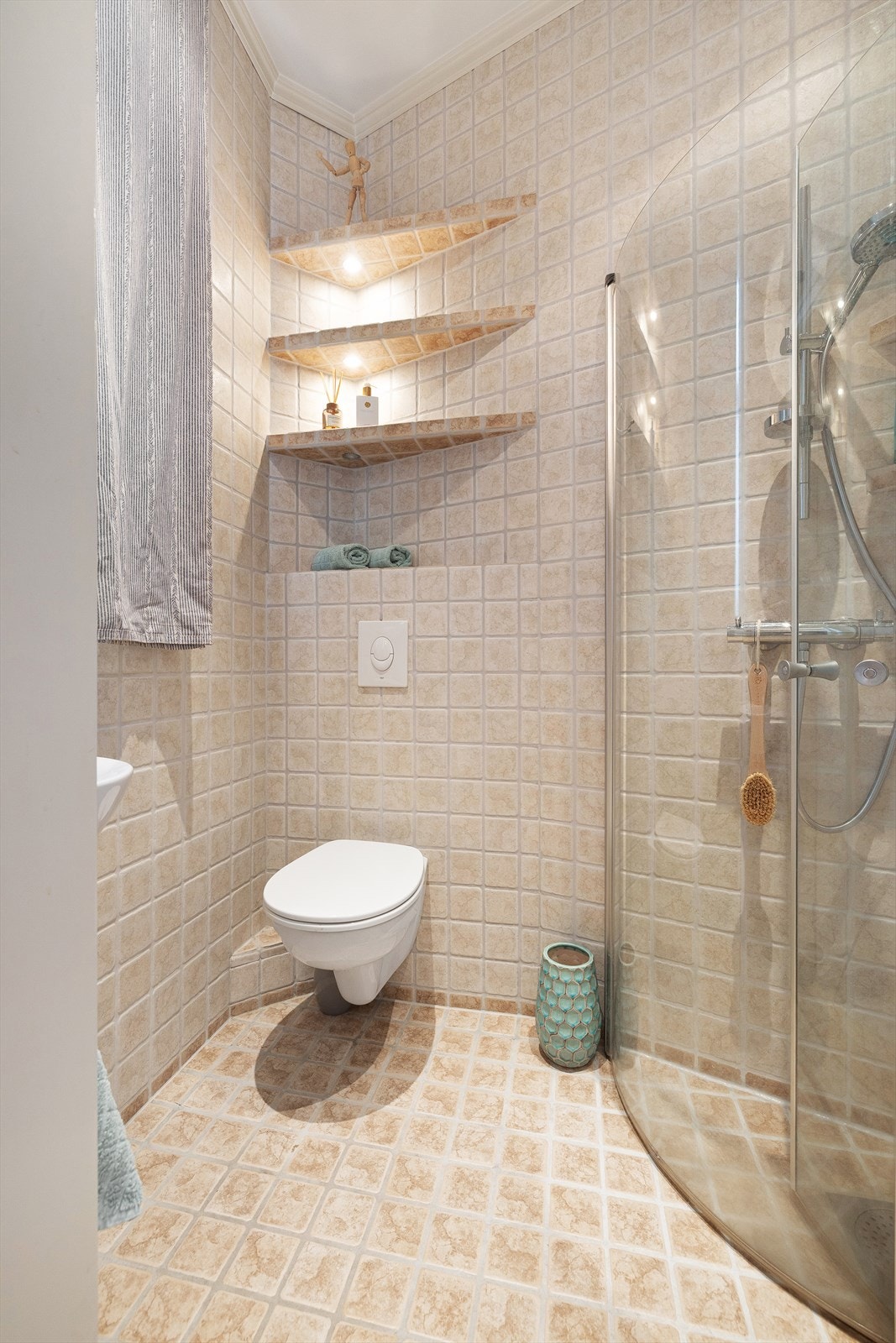 Dusjsone med innfellbare glassdører og dusjarmatur tilkoblet hånddusj, flislagte hyller på vegg med integrert belysning, og vegghengt toalett. Galleribilde