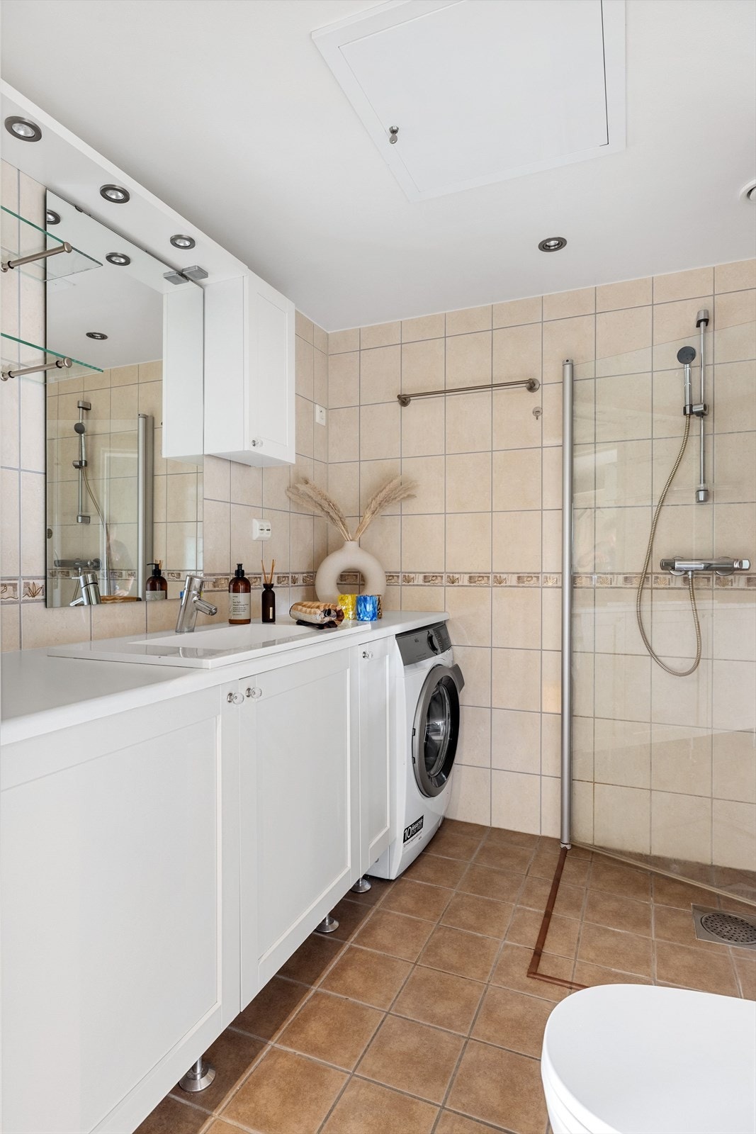 Lys baderomsinnredning med benkeplate og nedfelt servant, speil med belysning, glasshyller og vegghengt skap over. Praktisk med plass og opplegg for vaskemaskin under benkeplaten. Galleribilde