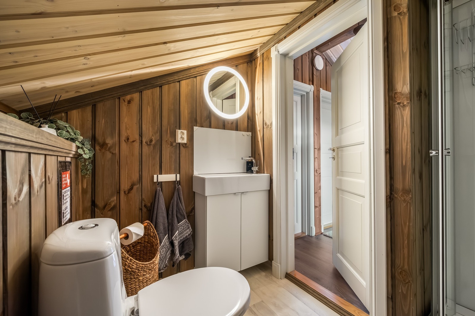 Gulvflater med gulvbelegg og veggflater med panel. Skråtak med panel. Dusjkabinett, gulvmontert toalett og servantskap med hvite slette fronter og heldekkende servant. Speil med integrert lys. Galleribilde