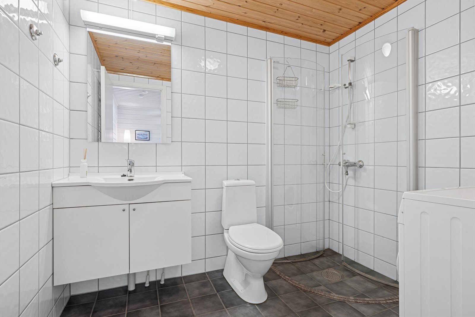 Helfliset baderom med innfellbare dusjvegger, servant med skap, gulvmontert toalett og opplegg til vaskemaskin. Galleribilde