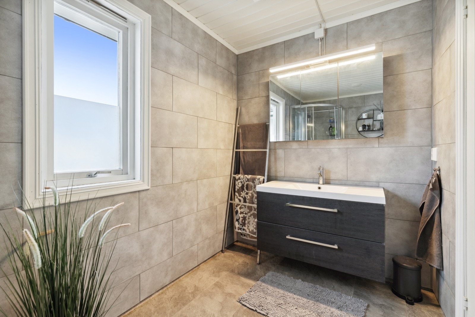 Eldre bad av ukjent årgang med vinylbelegg på gulv og baderomspanel på veggflater. Det er satt inn nyere baderomsmøblement med 2 skuffer og tilhørende speil. Galleribilde