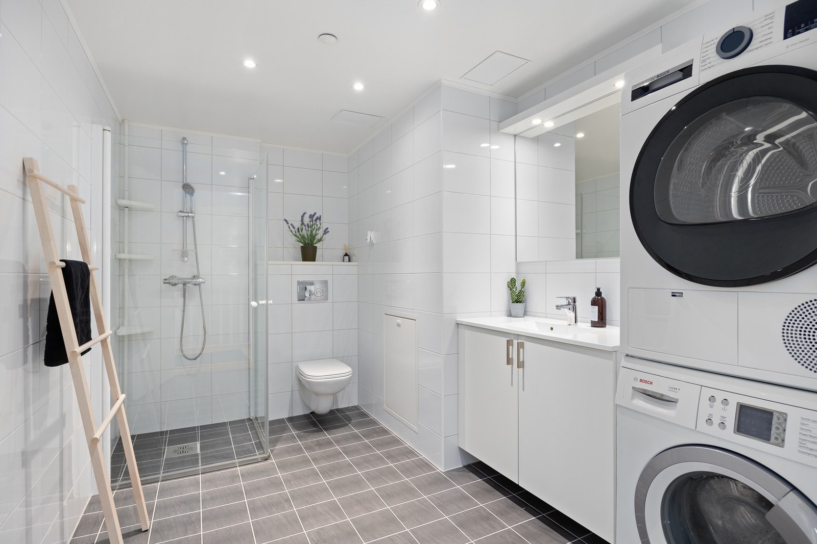 B A D E V Æ R E L S E II: Lyst og delikat badeværelse med vegghengte innfellbare dusjdører i herdet glass. Heldekkende servant med underskap, vegghengt speil med lys over Galleribilde