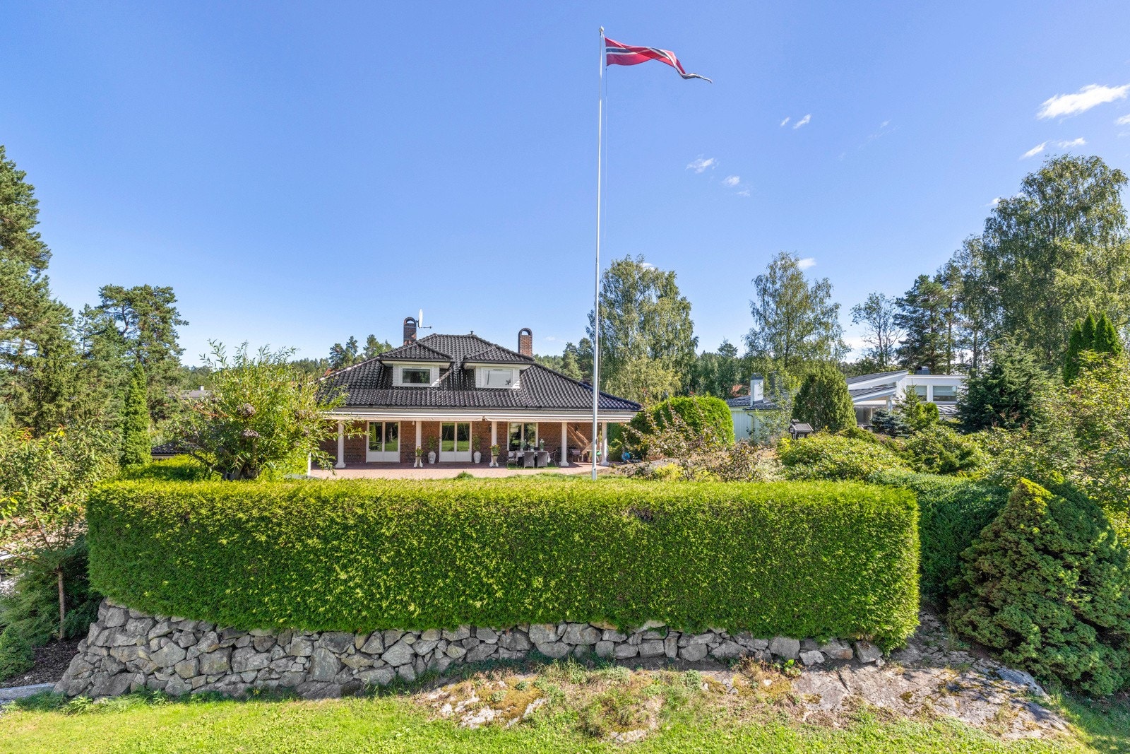 Eiendommen er innrammet av en frodig, grønn hekk og ligger skjermet til med gode solforhold. Flaggstangen i hagen setter den klassiske prikken over i-en.