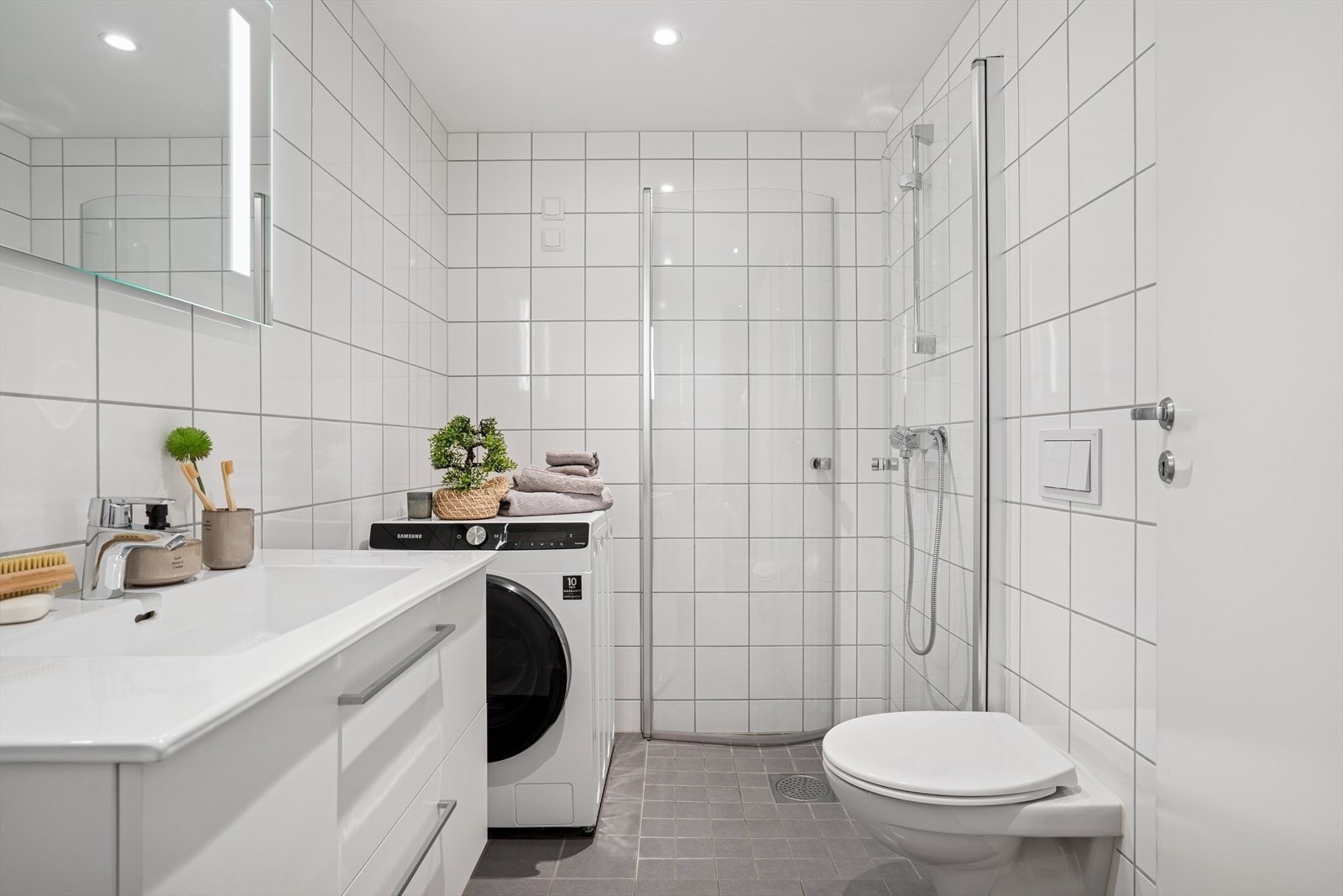 Leilighetens andre bad med et lekkert og stilrent utrykk og opplegg for vaskemaskin. Det er flislagt gulv med varmekabler. Galleribilde