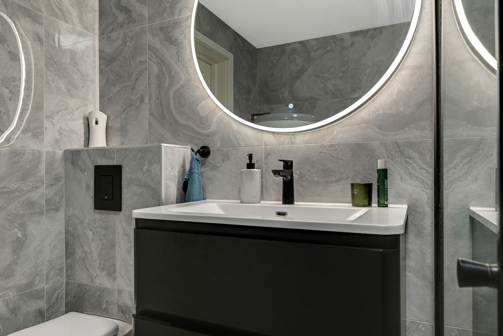 Badet inneholder et vegghengt speil med ringlys, en servant med underskuffer, et dusjhjørne med glassdører og et vegghengt klosett. I tillegg er det et vegghengt høyskap for ytterligere oppbevaring. Galleribilde