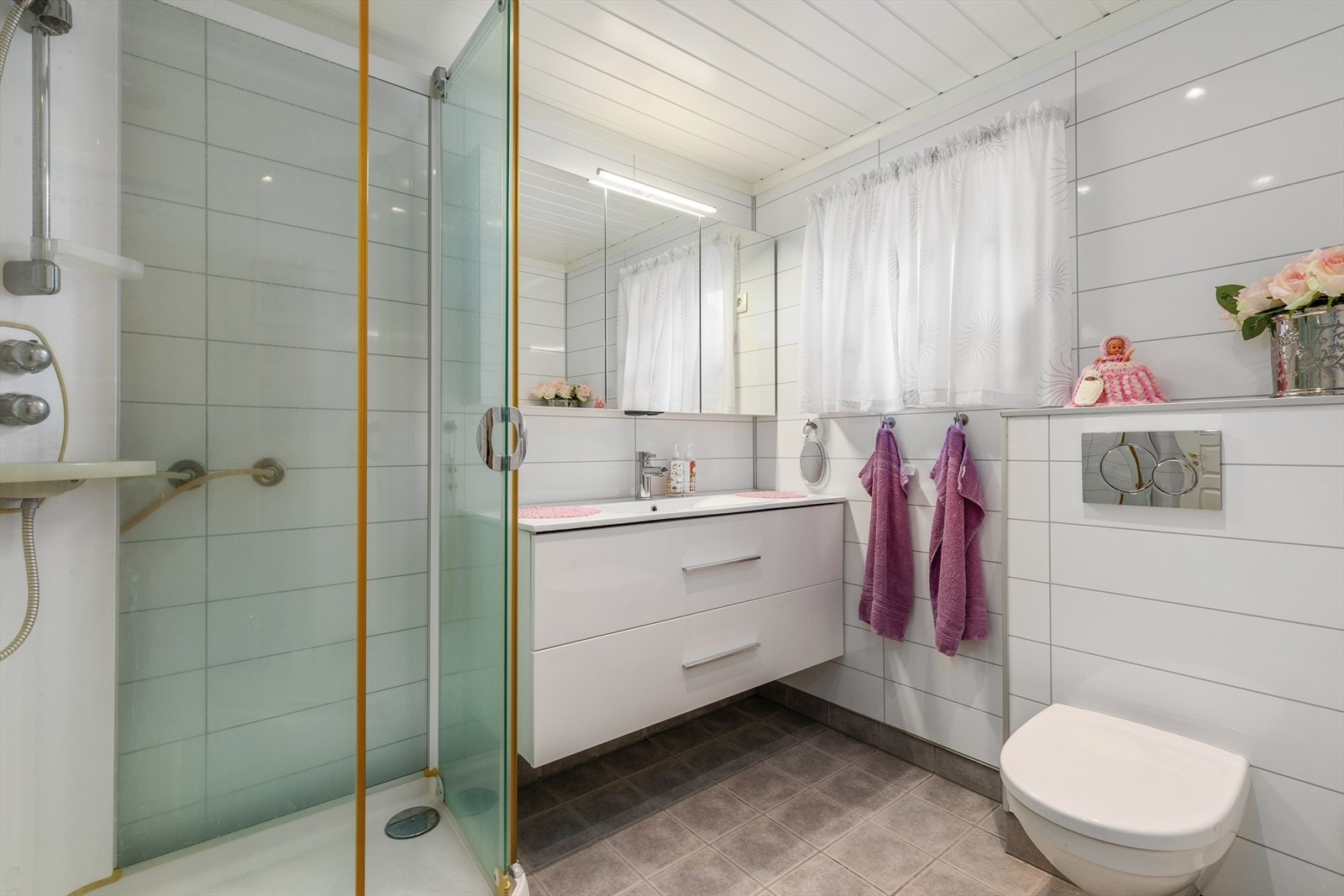 Bad med flislagt golv på eksisterande belegg, baderomsplater fra 2015 og plater i himling. Badet er utstyrt med dusjkabinett, baderomsinnredning med speilskap over og vegghengt toalett. Galleribilde