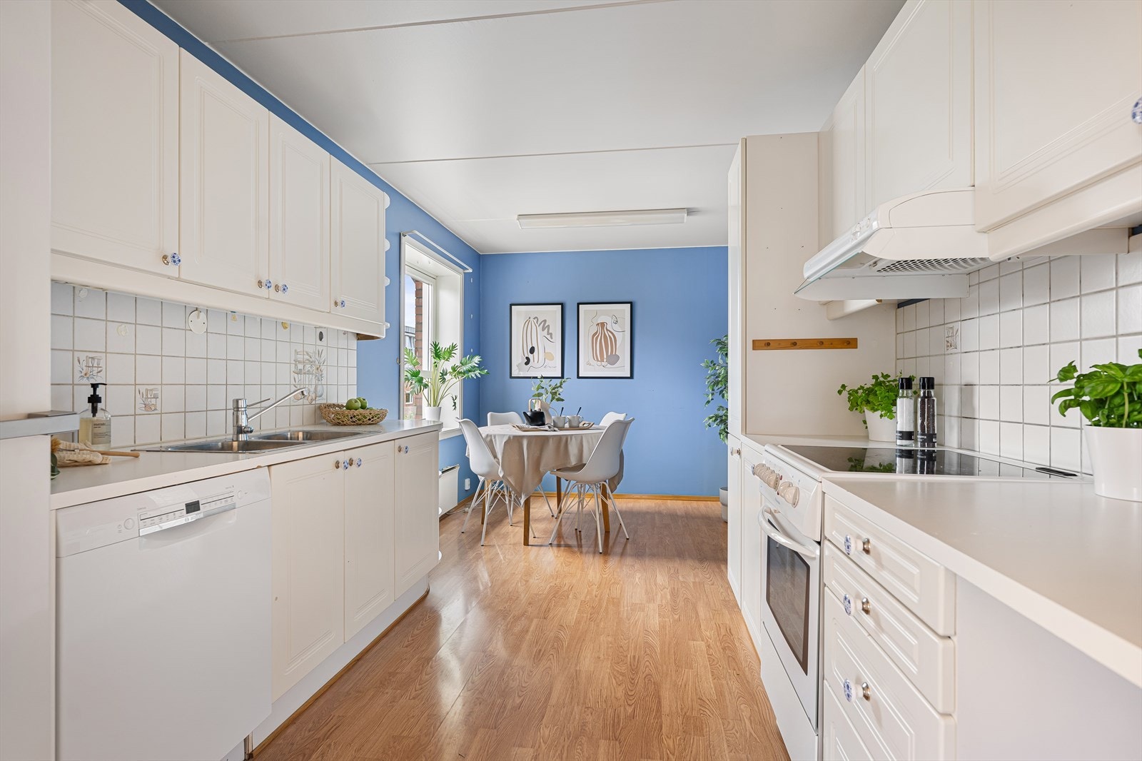 Kjøkken med eldre innredning bestående av hvite, profilerte fronter og 28 mm tykk hvit laminatbenkeplate med nedfelt dobbel vaskekum og ettgreps blandebatteri i rustfritt stål fra Mora. Galleribilde
