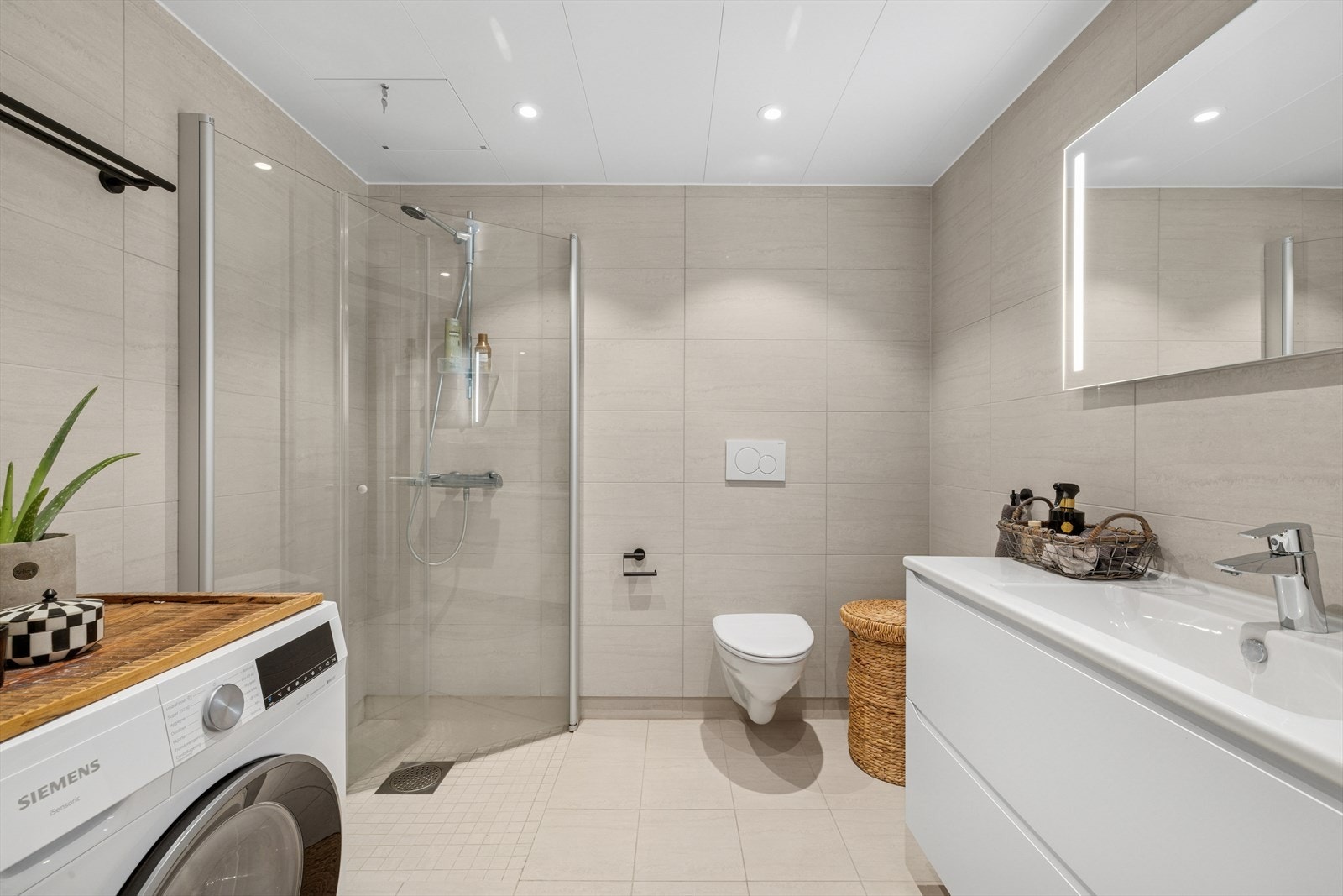 Moderne flislagt bad med dusjhjørne, vegghengt toalett, servantinnredning og opplegg for vaskemaskin. Galleribilde