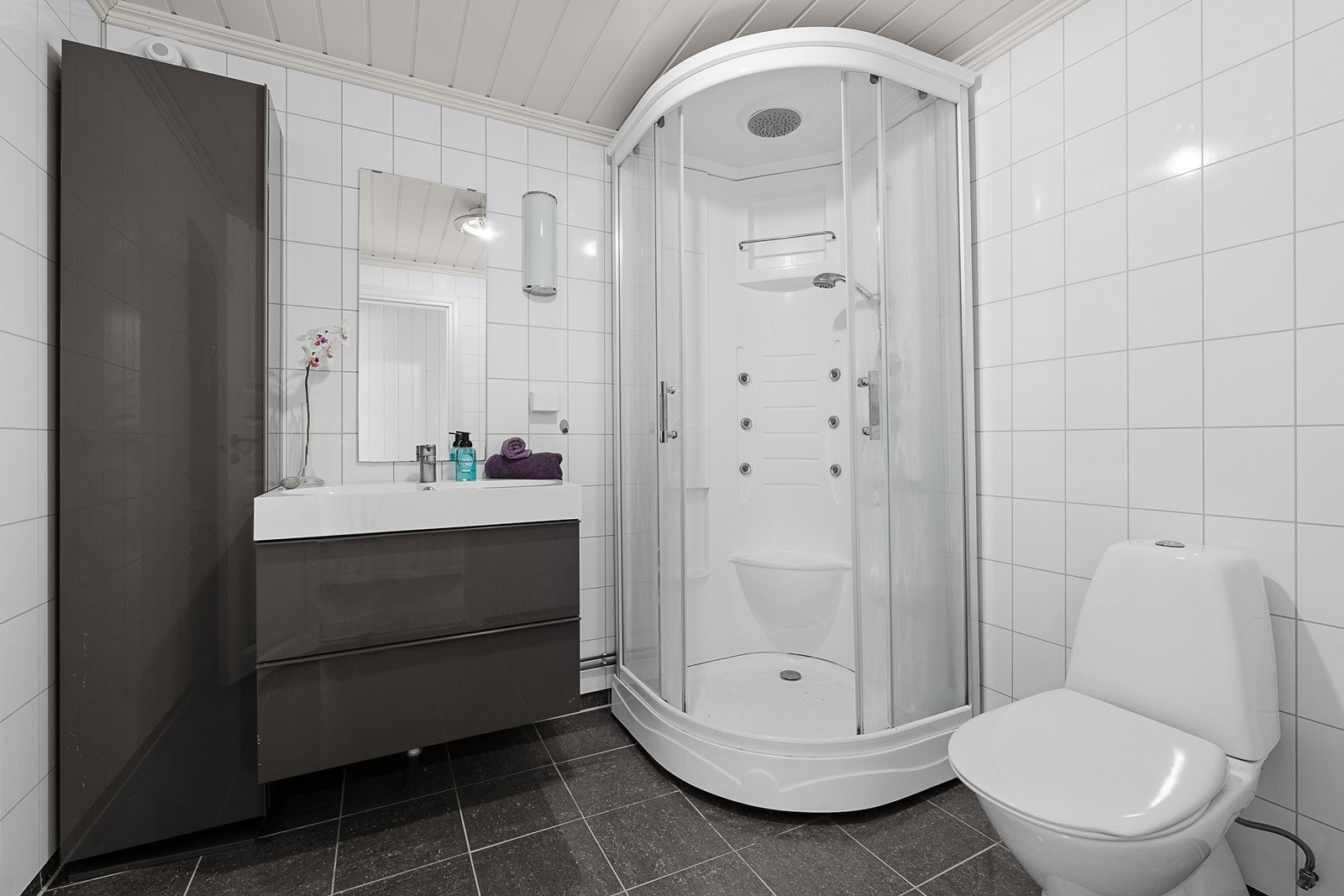 Badet er fra 2013 og har flislagt gulv med varmekabler og baderomsplater på vegger. Rommet inneholder dusjkabinett, servant med skuffer og skap og wc. Galleribilde