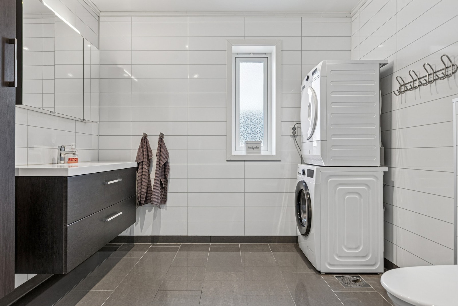 Badet i andre etasje fungerer også som vaskerom og har en moderne og plassbesparende løsning med opplegg for vaskemaskin. Rommet er utstyrt med servantskap, servant, dusjhjørne med innfellbare dører og vegghengt toalett. Galleribilde