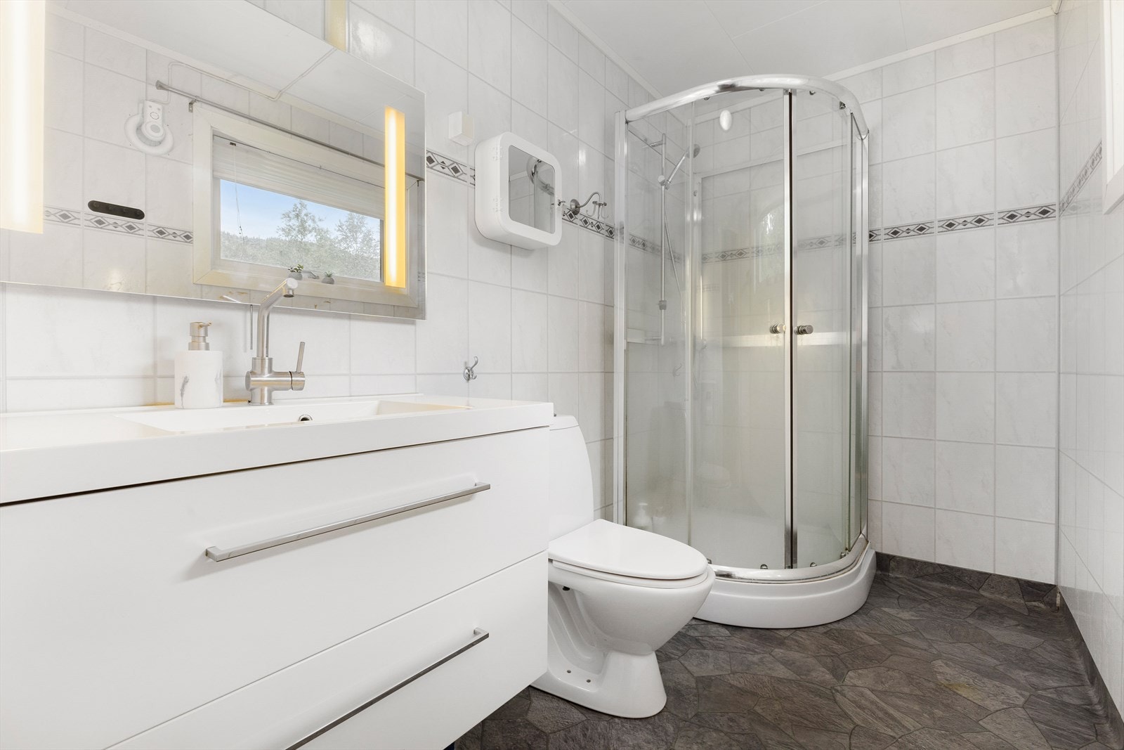 Badet ligger i direkte tilknytning gangen. Her er det innredet med dusjkabinett, toalett og en romslig baderomsinnredning. I tillegg er det plass til ekstra oppbevaring. Galleribilde