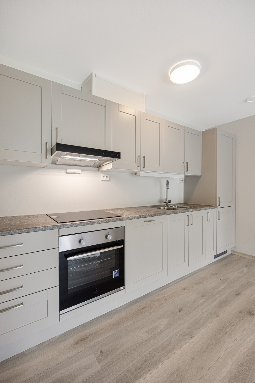 Innredningen har gråbeige rammefronter og en grå laminatbenkeplate med planlimt vaskekum i stål.
Hvitevarer som stekeovn, induksjonstopp, kombiskap og oppvaskmaskin er integrerte. Galleribilde