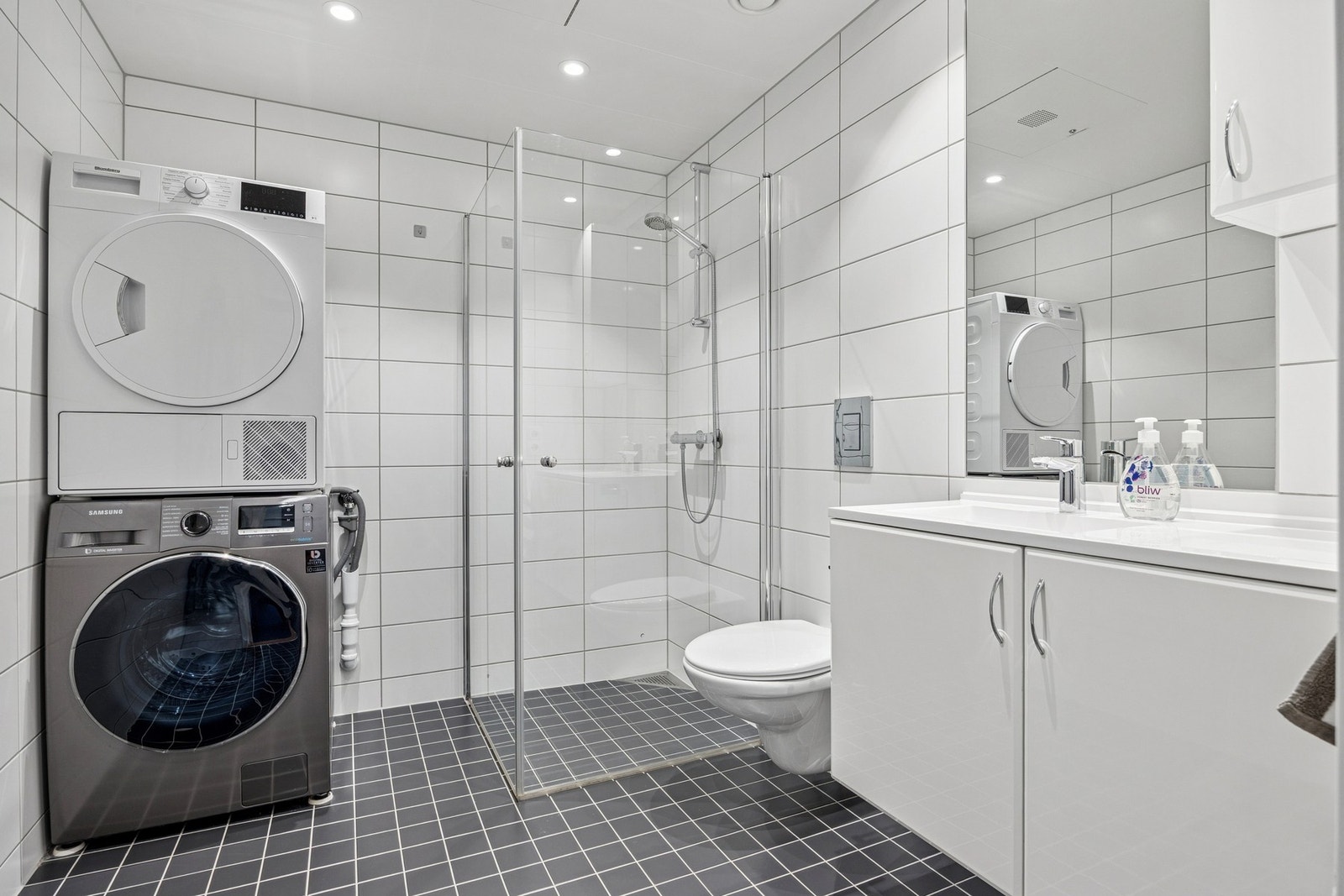 Lyst og tidløst baderom utstyrt med servantinnredning, speil, dusjhjørne, toalett og opplegg for vaskemaskin, samt downlights i himling. Galleribilde