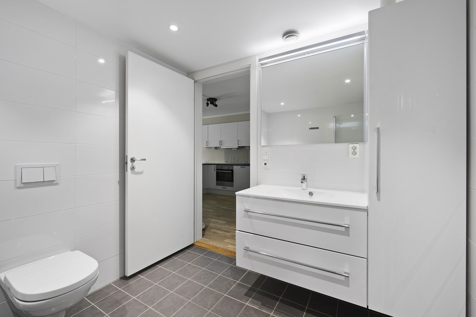 Delikat og lyst baderom innredet med vegghengt toalett, dusjnisje med glassdører, servant med underskap og sideskap, samt opplegg til vaskemaskin. Flis på gulv med varmekabler, flis på vegger og malte plater med downlights i himling Galleribilde