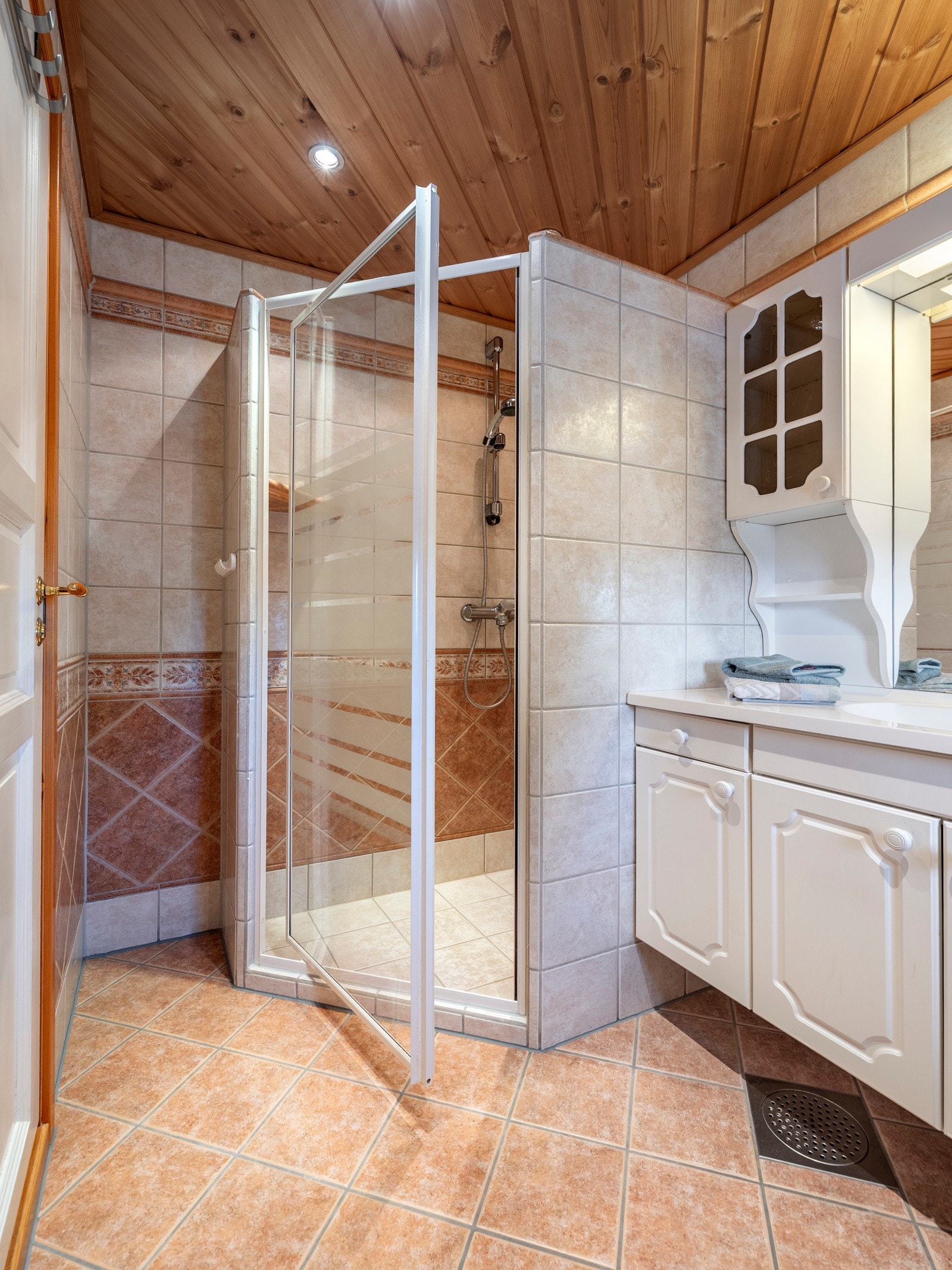 Badet er helfliset, og har elektrisk varme i gulvet, downlights i himlingen og et romslig dusjhjørne. Baderomsinnredningen har hvite, profilerte fronter og består av en heldekkende servant på en skapseksjon, to veggskap og et speil med belysning. Galleribilde