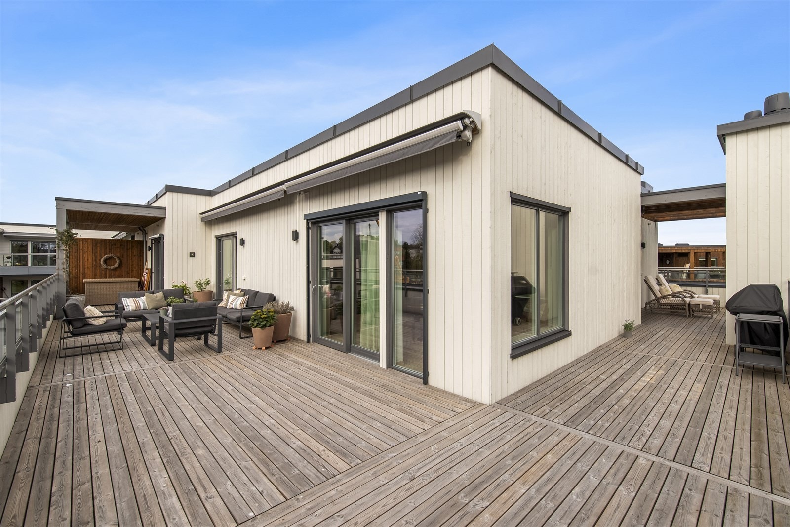 Mot vest vender terrassen ut mot grønne fellesarealer med gressplen og lekeplass. Terrassen har tett rekkverk med glass øverst, dekke av terrassebord og to elektriske markiser på vestsiden. Galleribilde