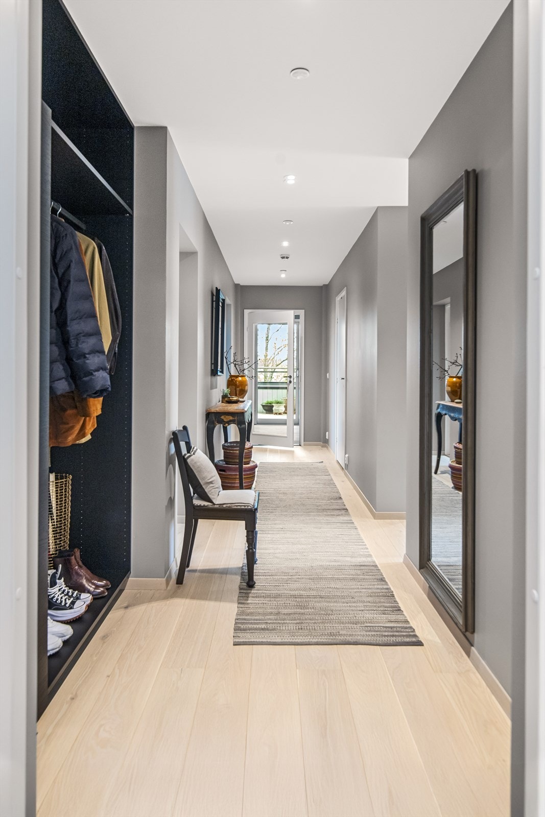 Ved inngangen er det en garderobenisje med mørk, takhøy garderobeinnredning, skap, hyller og garderobestang. I passasjen inn til stuen er det to boder, skjult bak skyvedører med speilfronter på den ene siden og lys front vis-à-vis. Galleribilde