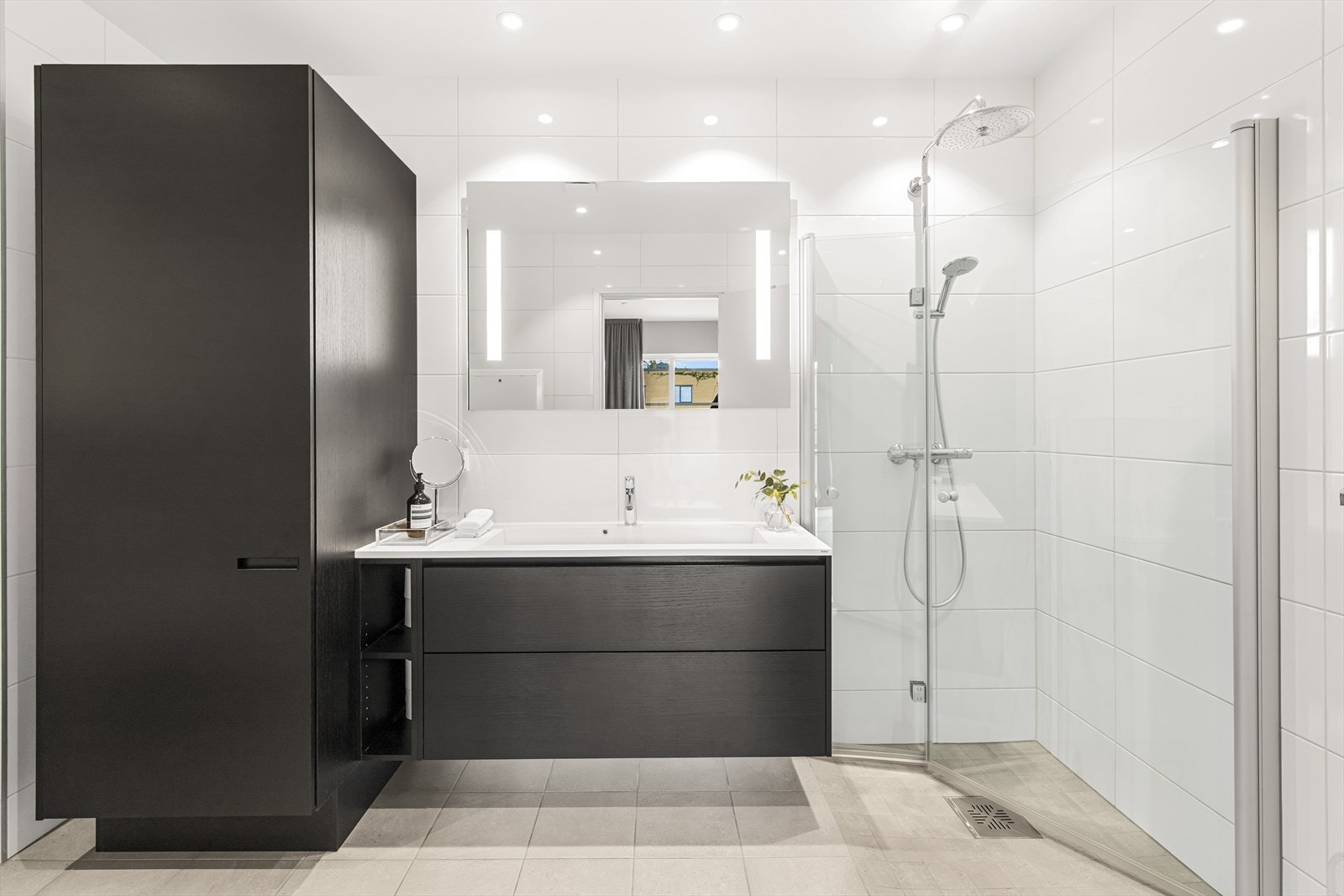 Begge har heldekkende servantplate (Kuma) på skuffeseksjon med sorte fronter i trestruktur, speil med integrert belysning, veggmontert toalett, og et dusjhjørne med innfellbare glassdører, regnfallsdusj og hånddusj. Galleribilde