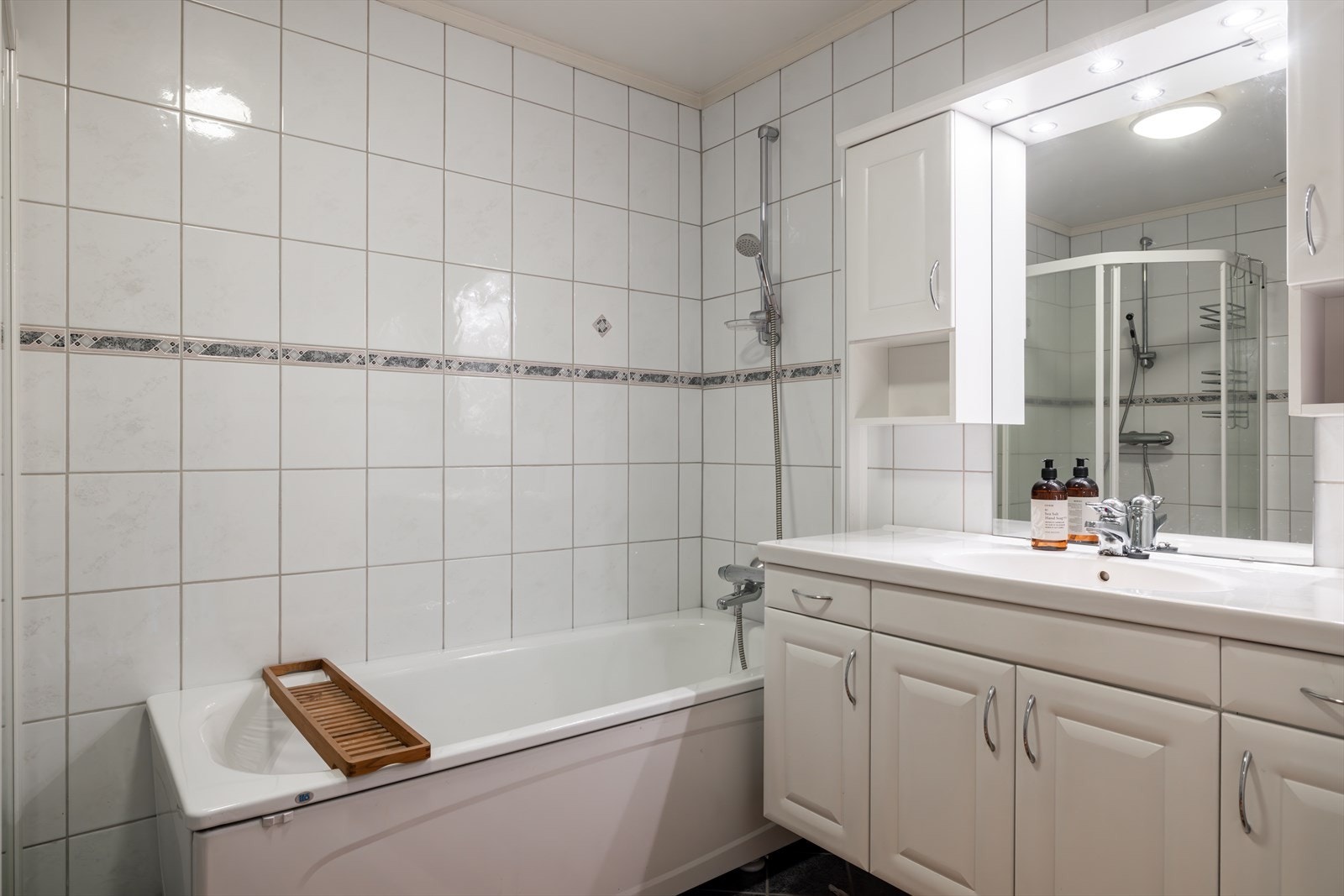 Innredningen består av en bred, heldekkende servantplate på hvite servantskap, to veggskap, speil med belysning og toalett. Badet har mørke gulvfliser, hvite veggfliser og varmekabler i gulvet. Galleribilde