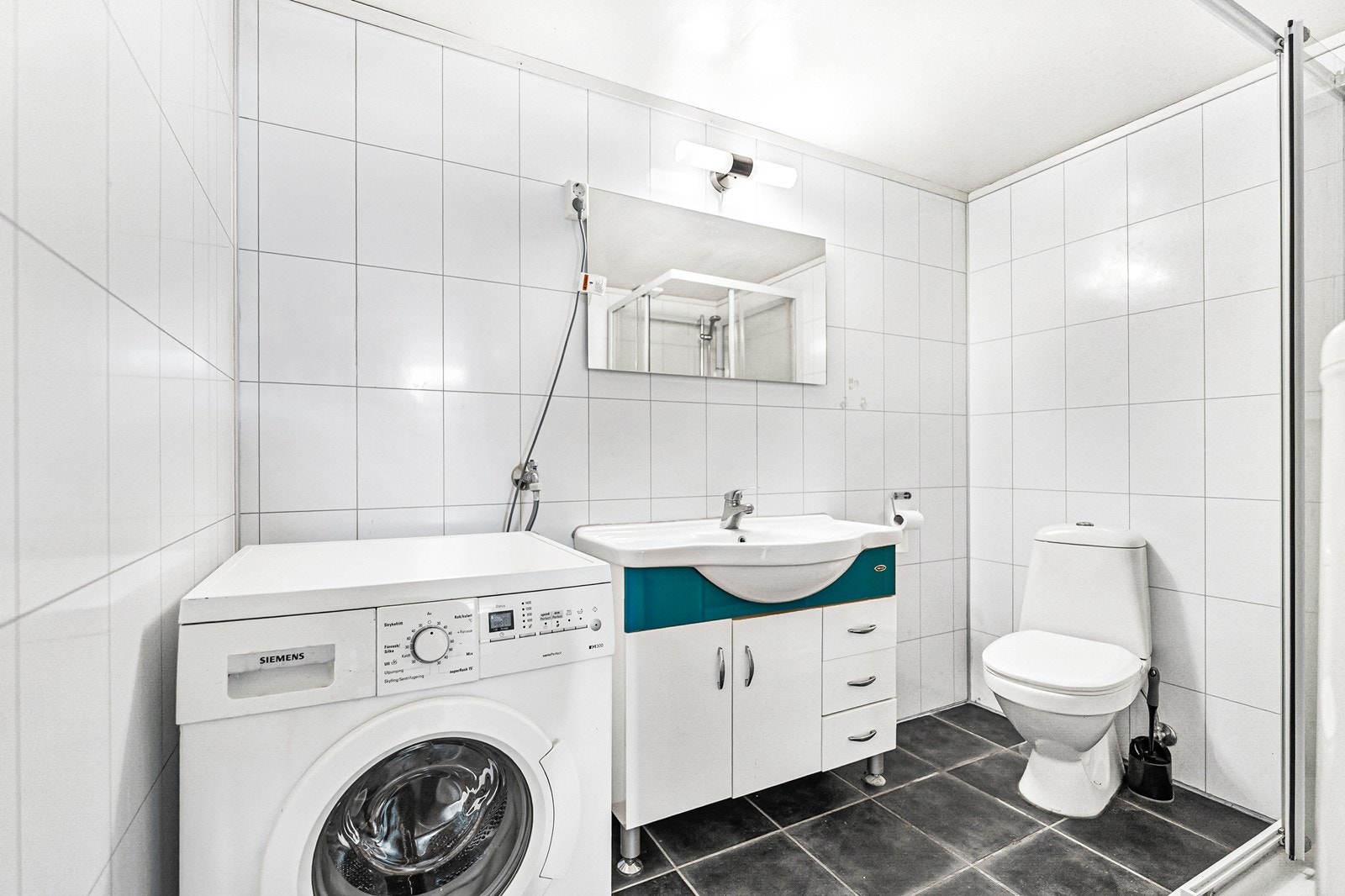 Badet er innredet med toalett, speil, dusjkabinett, servant med skap og opplegg for vaskemaskin. Galleribilde