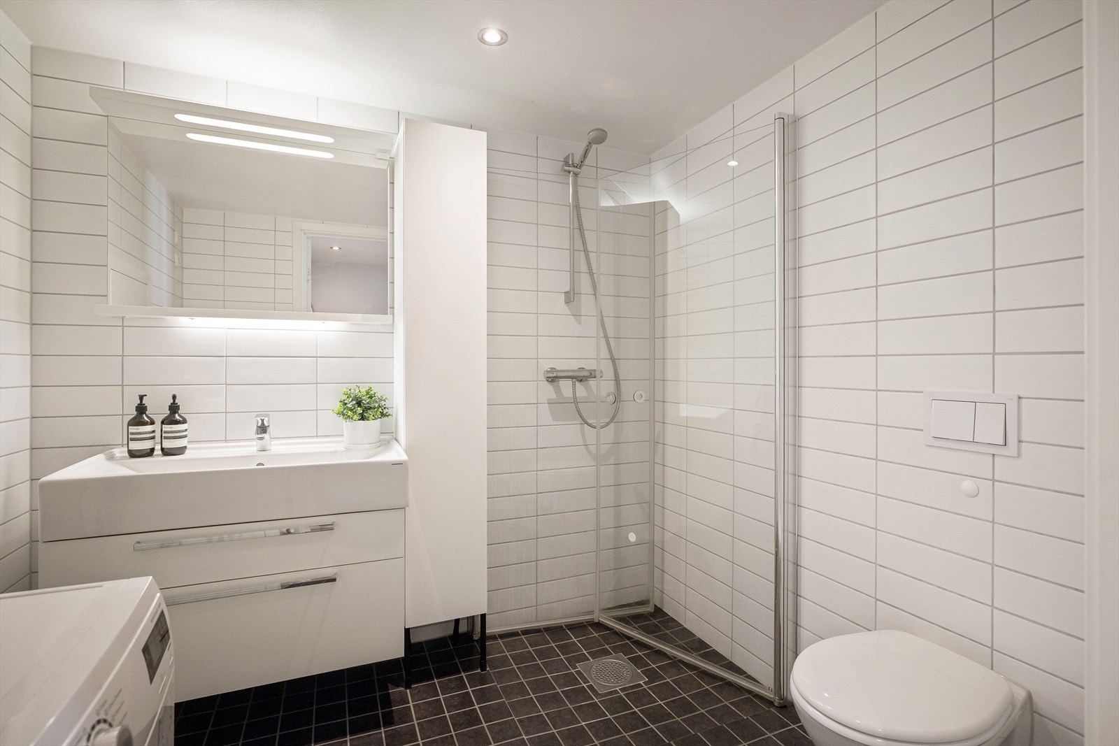 Badet er utstyrt med veggmontert toalett, dusjhjørne med innfellbare dusjvegger i glass og baderomsinnredning med skuffer, servant med ettgreps blandebatteri fra HansGrohe og speil med belysning. Galleribilde