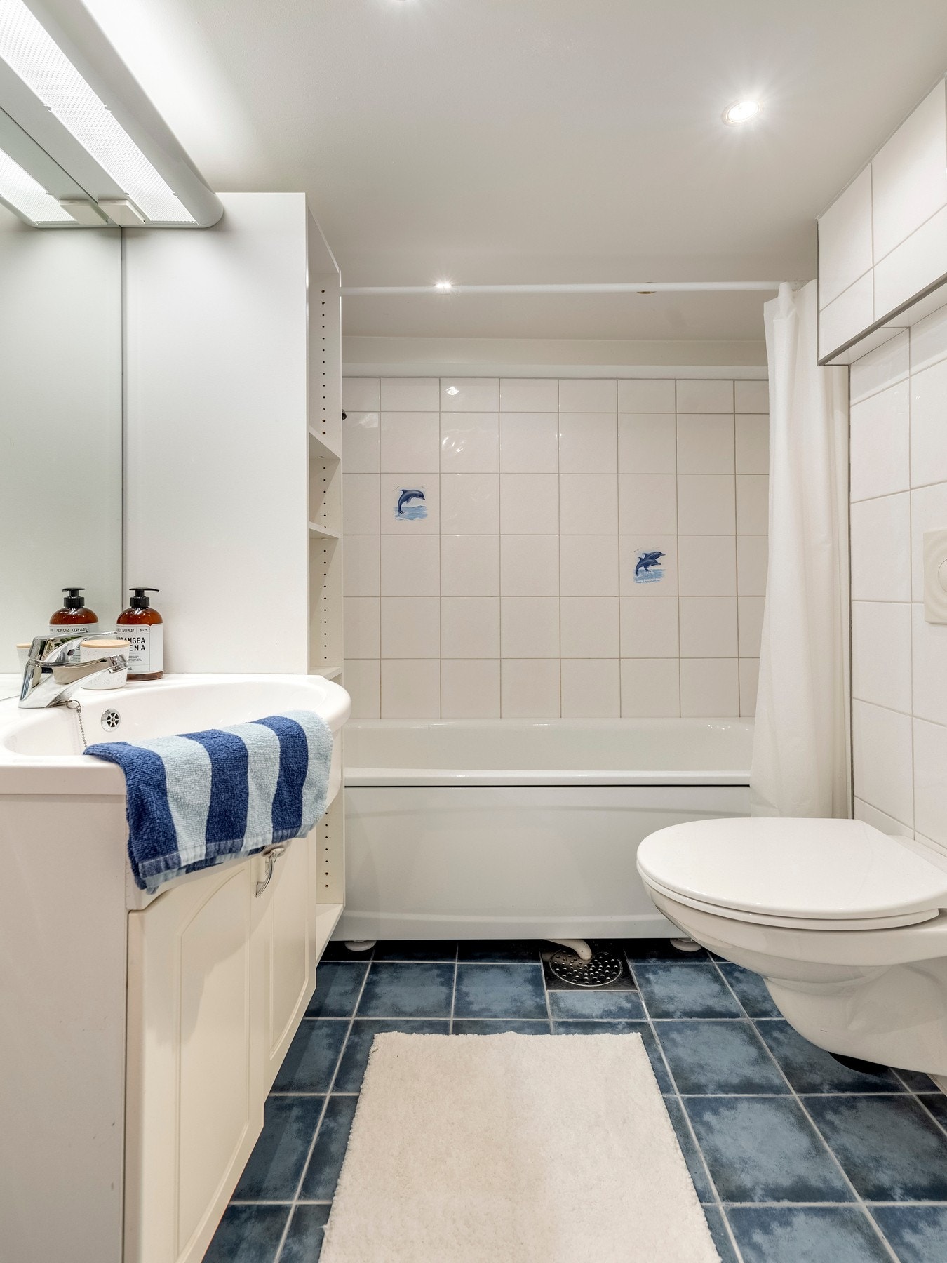 Badet er utstyrt med servantinnredning, speil på vegg over servant, ett høyskap, vegghengt toalett og badekar. Galleribilde