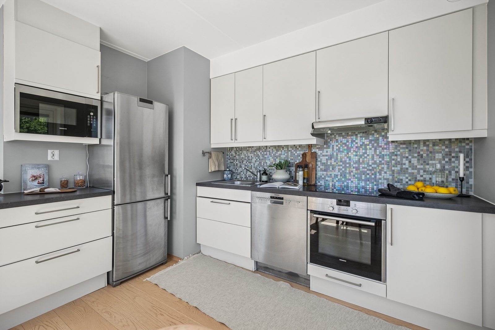 Kjøkkenet har en stilren innredning med glatte fronter, elegant belysning under overskapene og delikate fliser på veggen over benkeplatene. Galleribilde