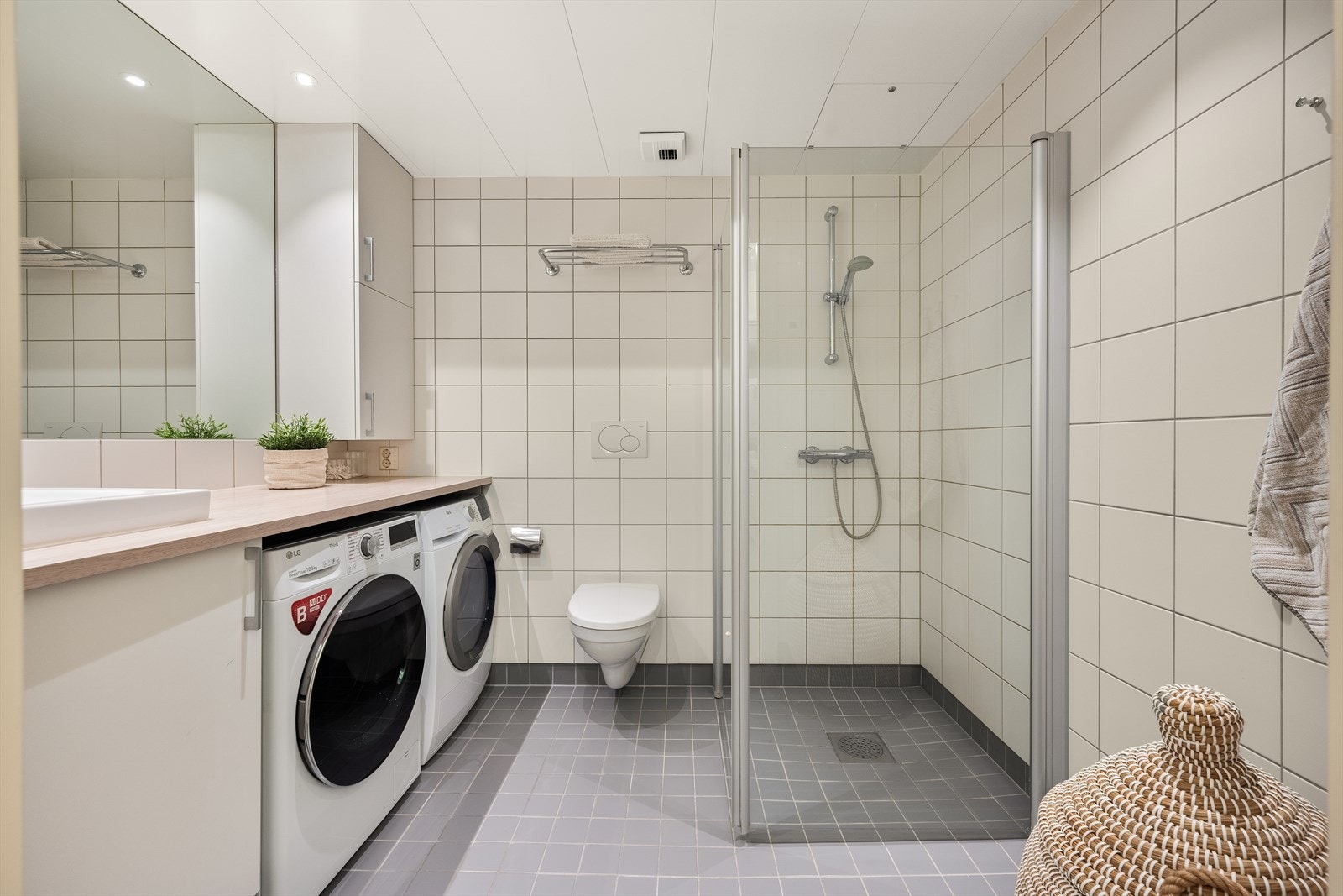 Badet er innredet med vegghengt toalett, dusjhjørne med svingbare glassdører og bred benkeplate med nedfelt servant, skap og speil. Galleribilde