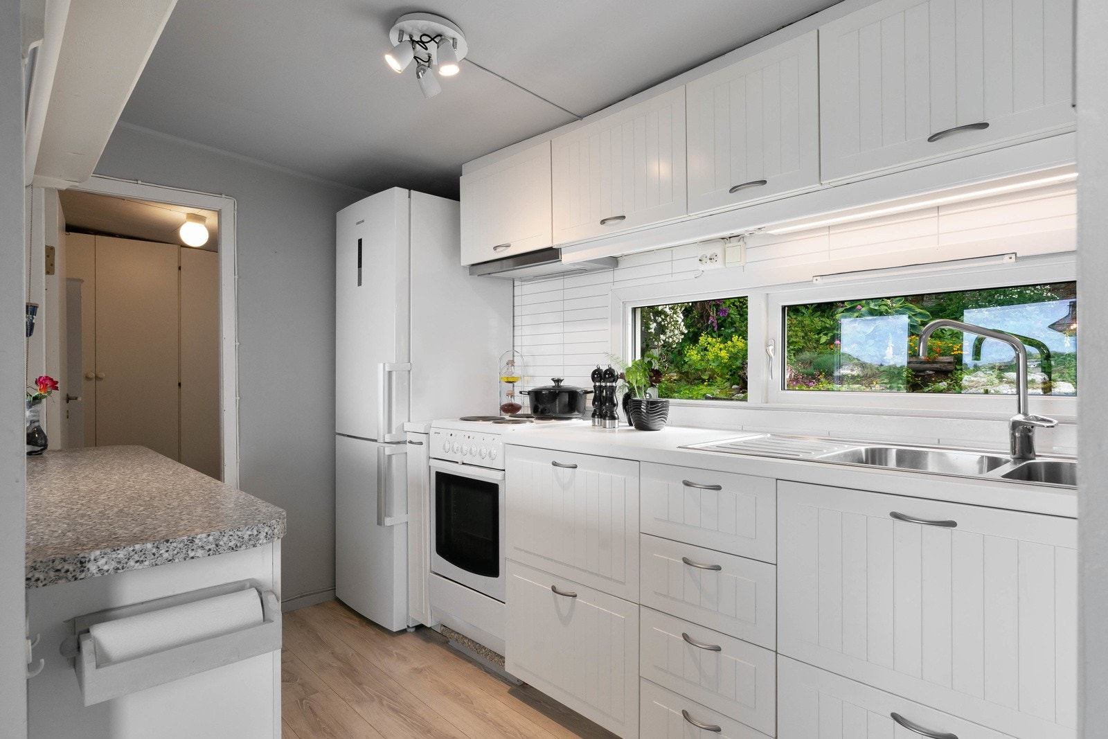 Kjøkkeninnredningen er montert langs to vegger og har hvite, profilerte fronter og benkeplate i laminat. Det er nedfelt oppvaskkum med avrenningsfelt i den ene benken. Galleribilde