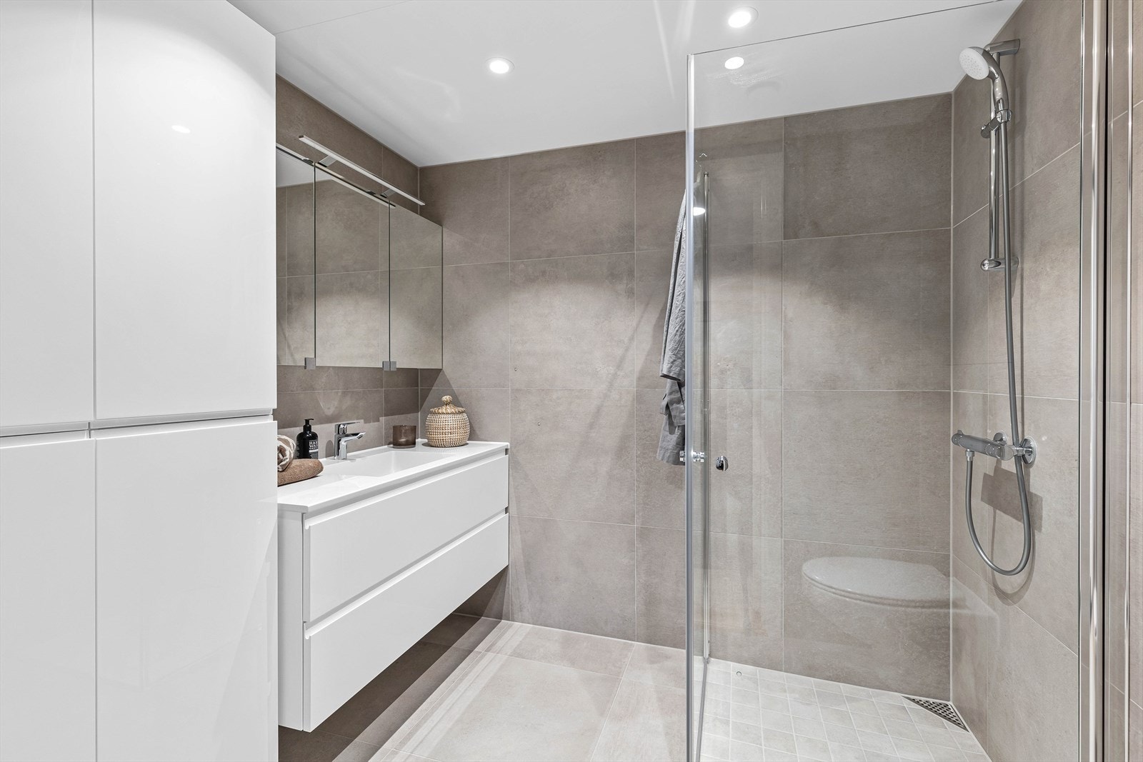 Badet i tilknytning til mastersoverommet er av moderne stil- og fargevalg. Det er innredet med innfellbare dusjdører, veggmontert toalett- og servant med oppbevaringsskuffer. Du får også godt med lagringsplass i veggmonterte skap og overskap med speil. Galleribilde