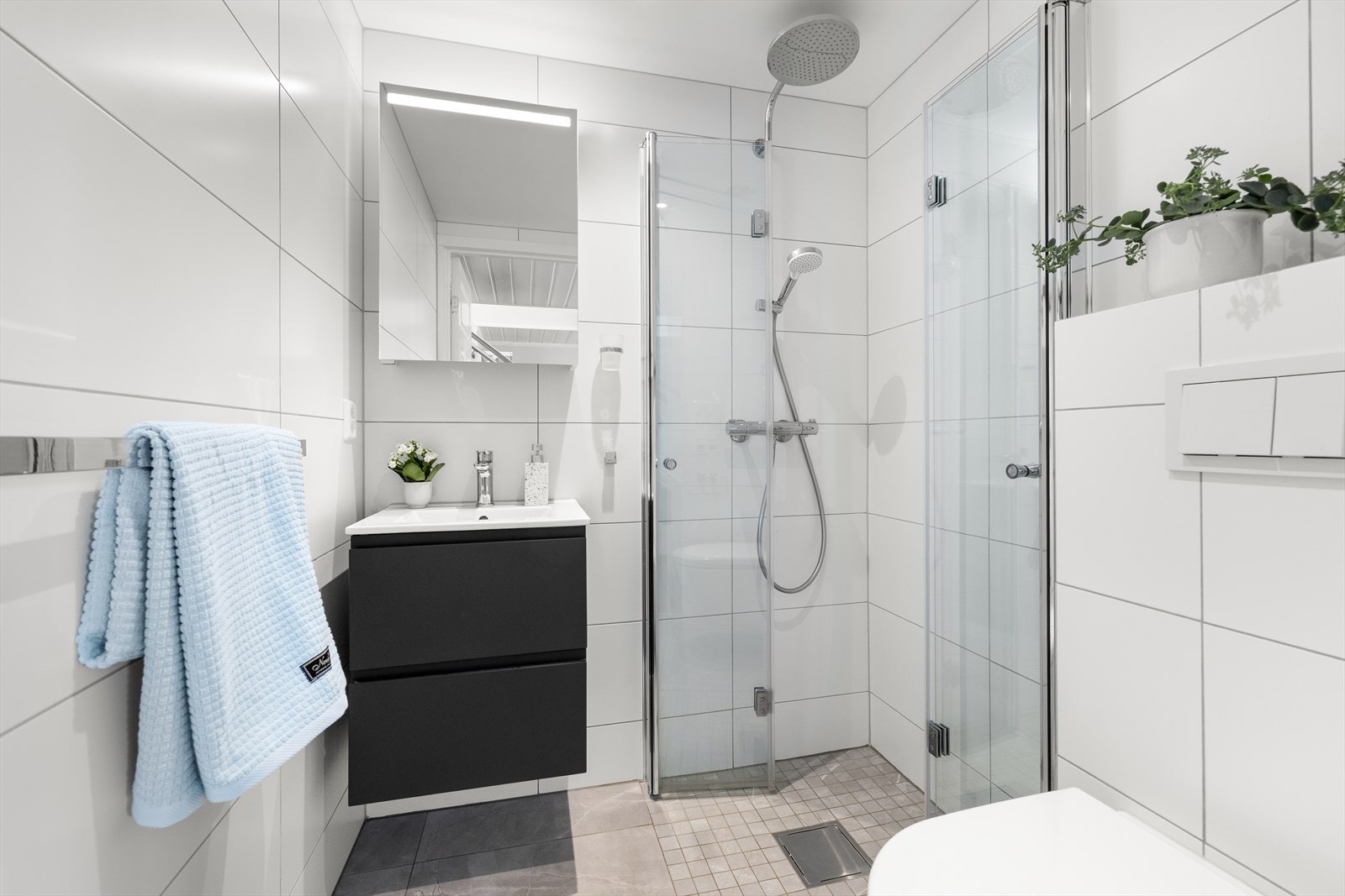 Pent flislagt baderom i tidløse fargetoner. Badet er utstyrt med vegghengt toalett, servant med skuffer og dusjnisje med innfellbare glassdører. Galleribilde