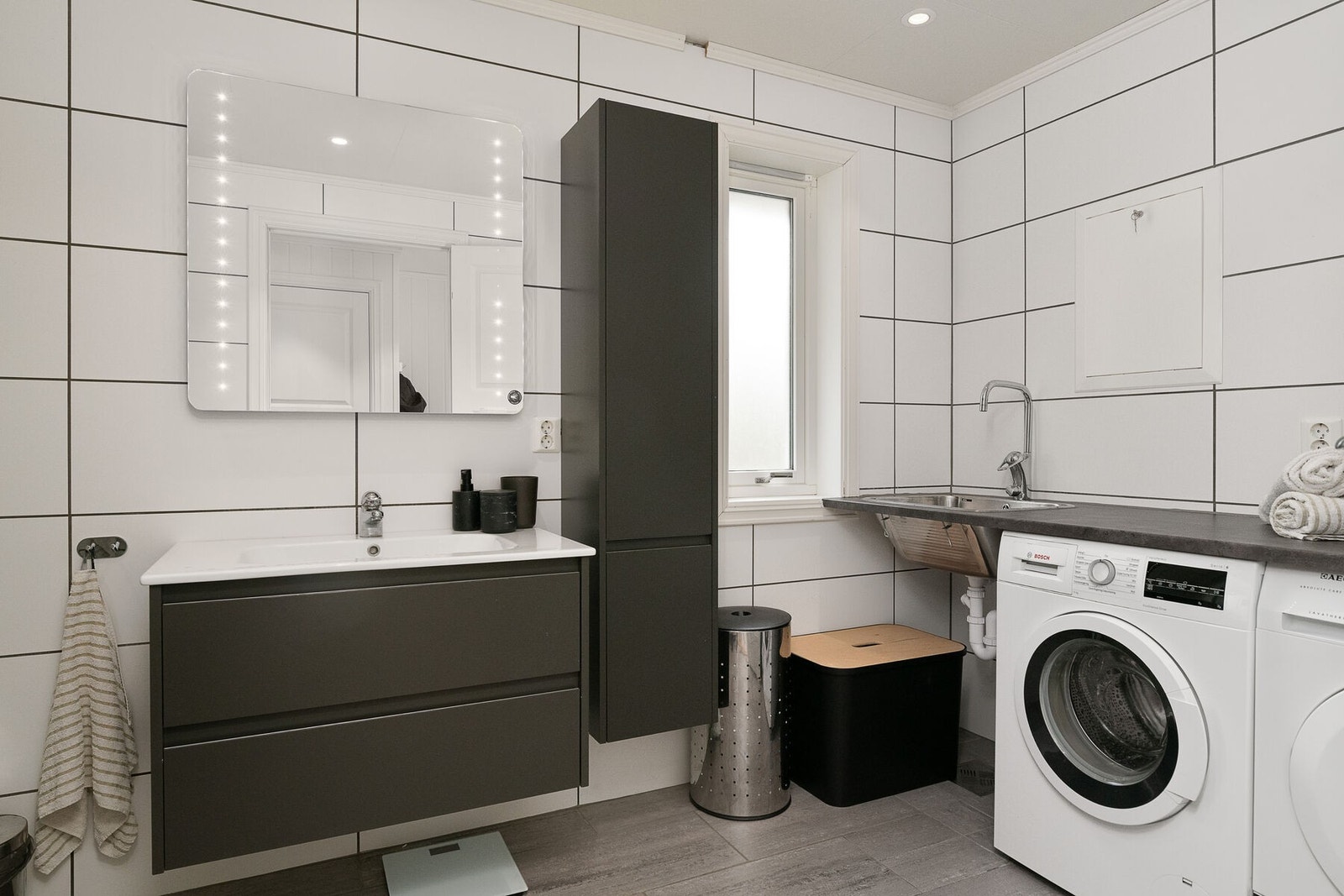 Bad/vaskerom har servant på innredning, veggmontert toalett, dusjnisje med skyvedør, benkeplate med skyllekar og opplegg for vaskemaskin Galleribilde