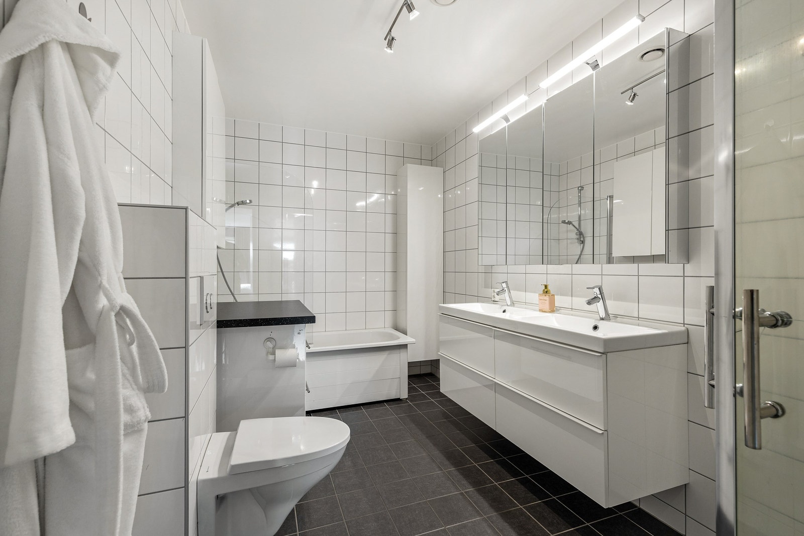 Speilskap med overlys over servant. Vegghengt baderomsmøbel med skuffer. Badekar med vegghengt badekararmatur. Vegghengt toalett. Galleribilde