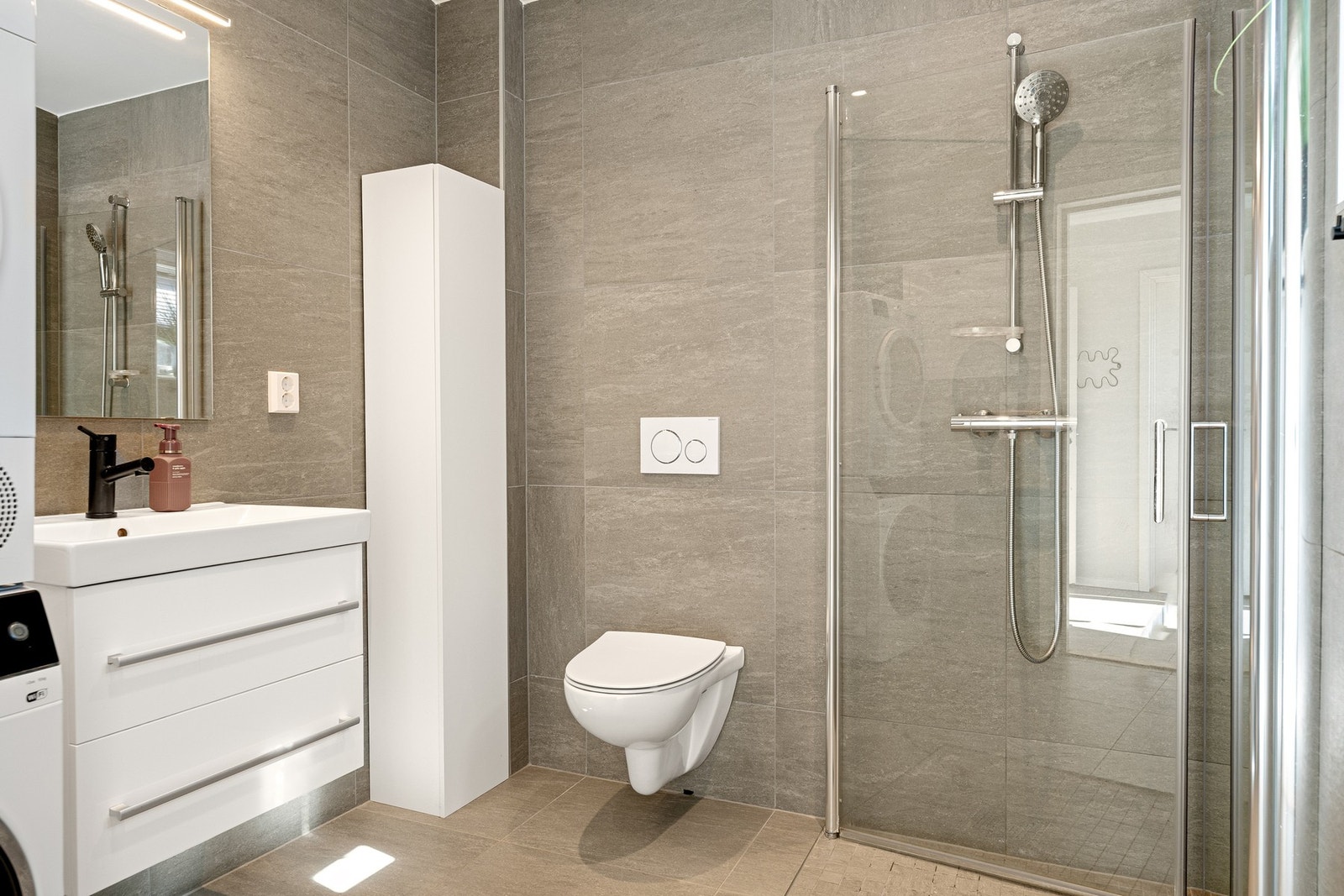 Lekkert flislagt bad/vaskerom med innredning, dusjhjørne, vegghengt toalett og opplegg for vaskemaskin. Galleribilde