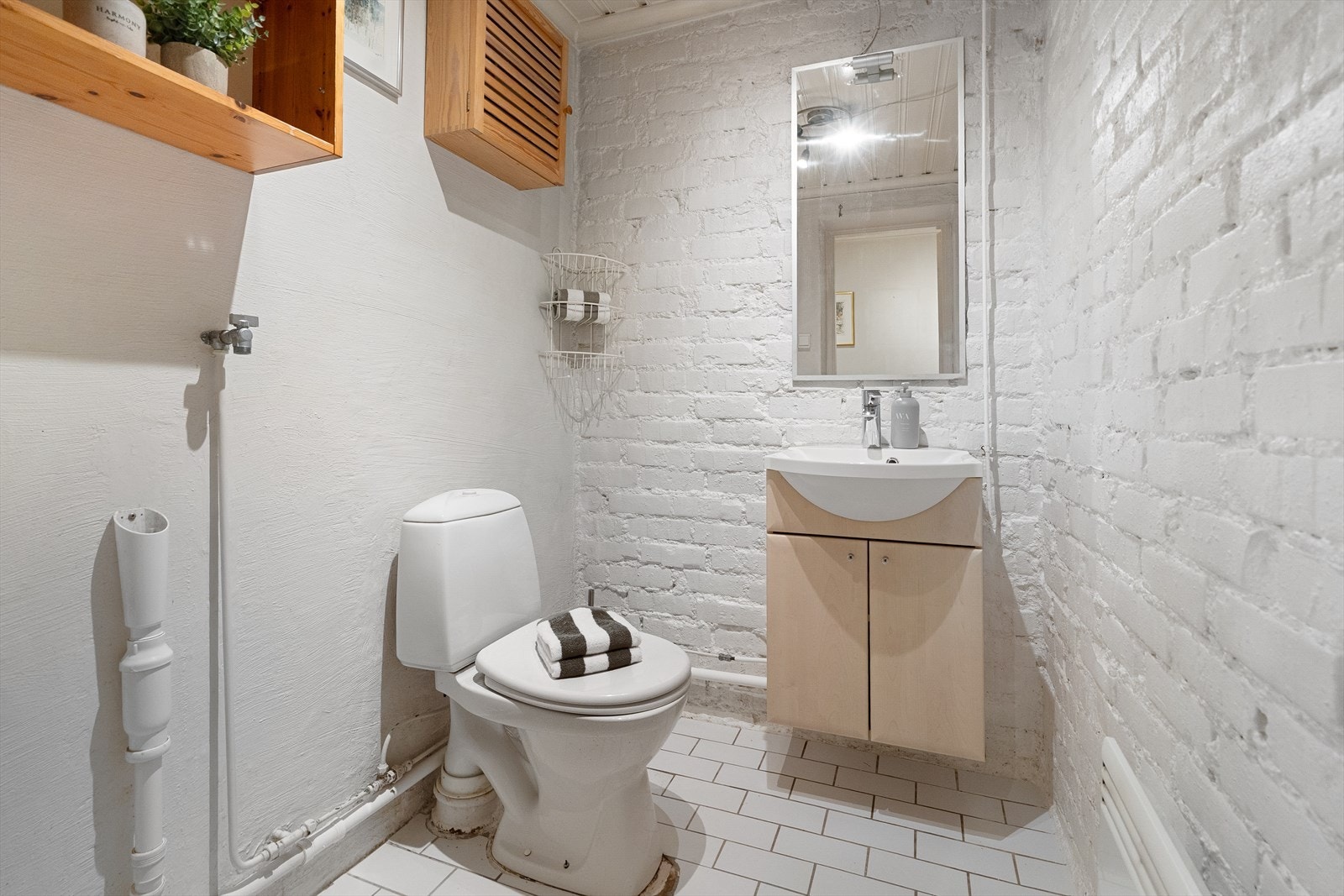 Eldre toalettrom med flis på gulv og pusset overflate på vegger og malt teglstein. Rommet er utstyrt med gulvmontert toalett, servantskap med hvitvservant og blandebatteri, opplegg for vaskemaskin. Galleribilde