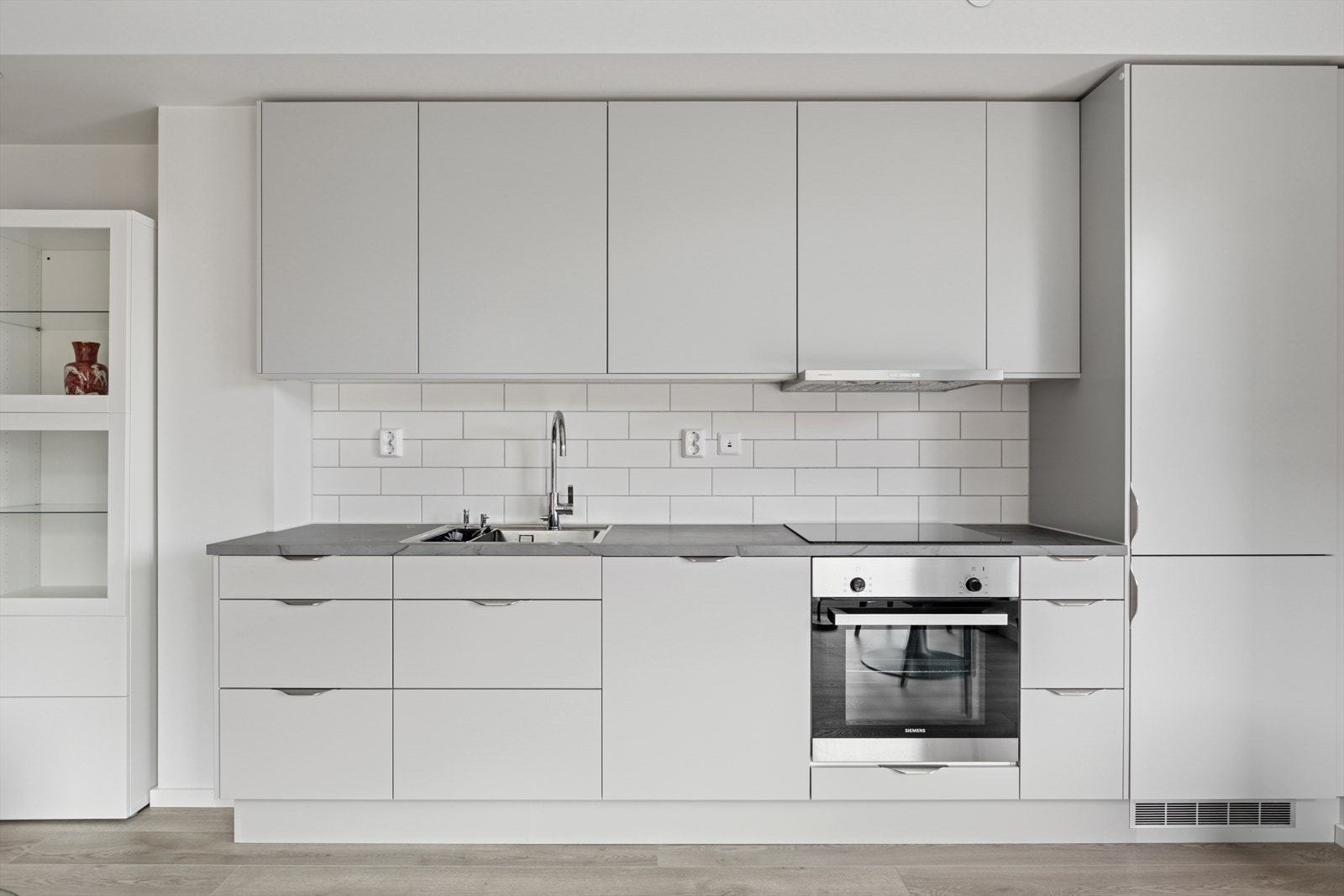 Innredningen har malte glatte fronter, laminat benkeplate, fliser over benken, ventilator med stål hette og stål benkebeslag. Galleribilde