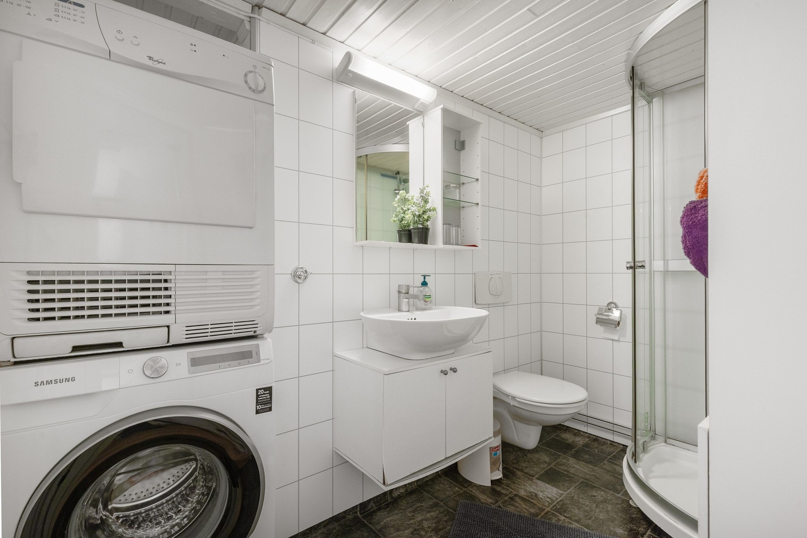 Baderommet er bes3tående av servant med underskap, vegghengt toalett, dusjkabinett og opplegg fro vaskemaskin og tørketrommel Galleribilde