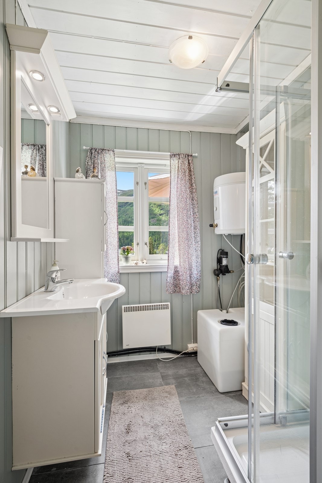 Badet har ikke innlagt vann, men dusj og servant får vanntilførsel fra en vannbeholder. Innredning med dusjkabinett, servantskap med underliggende oppbevaring, høyskap for ekstra lagringsplass, veggmontert speil og belysning. Nytt gulv i 2022. Galleribilde