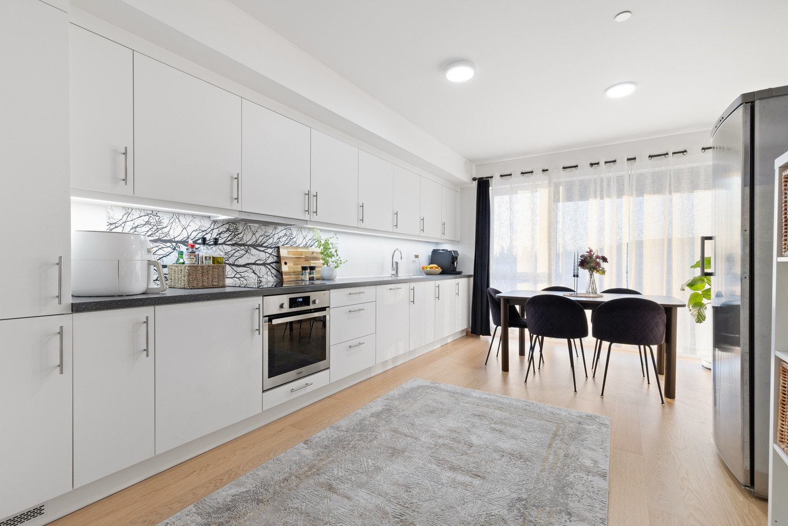 Stilrent kjøkken fra Hettich i åpen løsning mot stuen. Innredningen består av hvite slette fronter med foring opp til tak og laminert benkeplate i mørk utførelse. Nedfelt vaskekum i rustfritt stål med ett-greps blandebatteri. Galleribilde