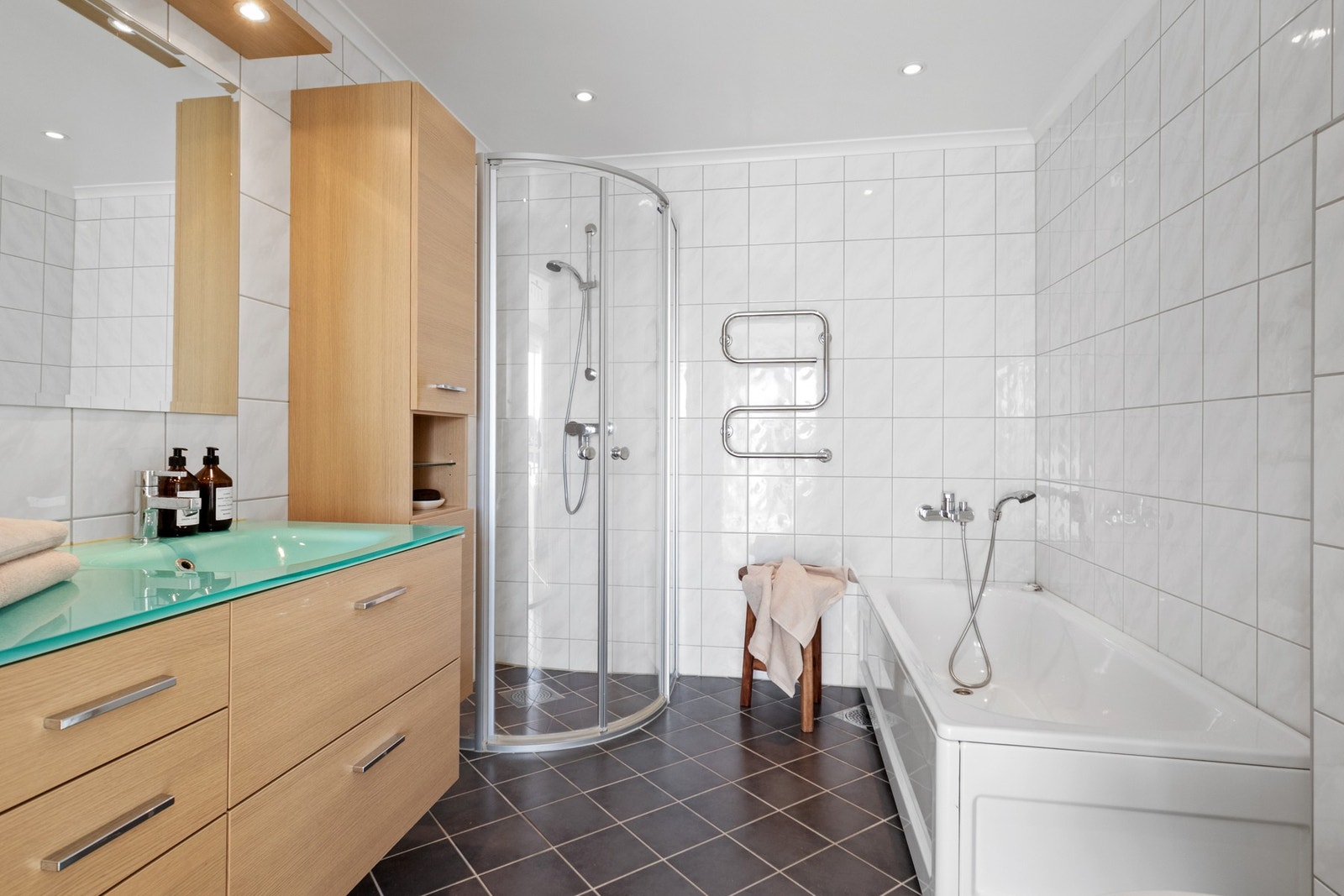 Baderomsinnredning med heldekkende servant, skuffer og speil med belysning. Dusjhjørne med glassdører, badekar og vegghengt toalett. Galleribilde