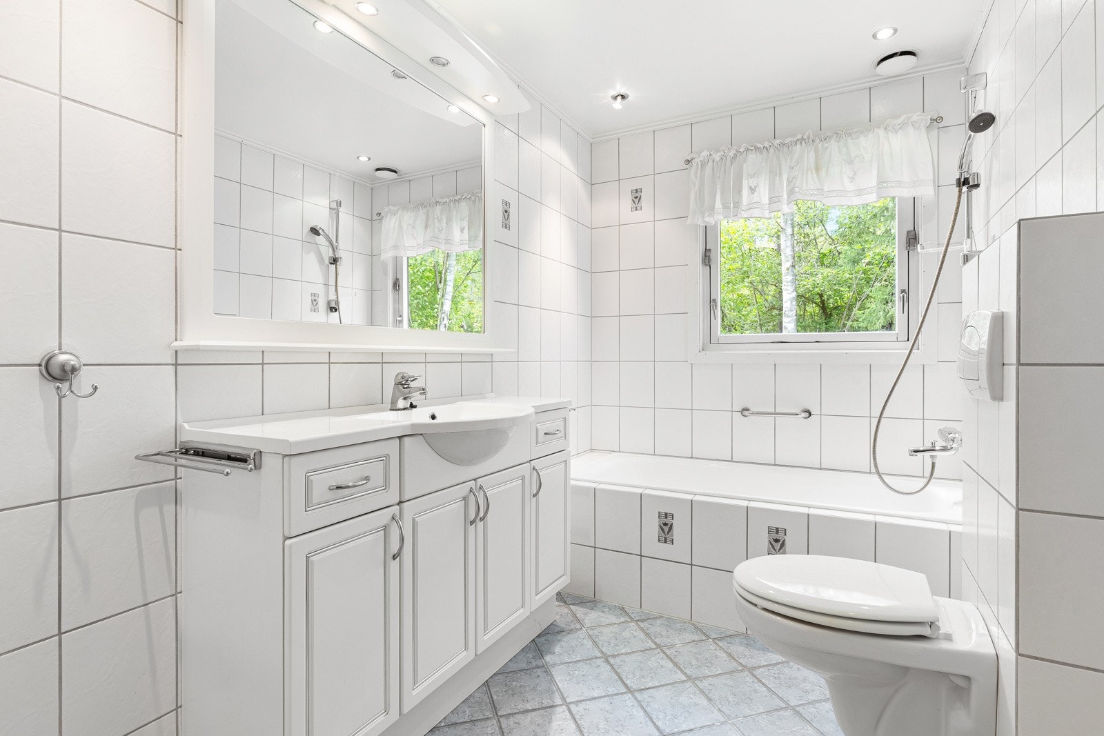 Flislagt gulv med gulvvarme. Flislagte vegger. Himling i lysmalt slett flate med downlights. Vegghengt servantinnredning med glatte lyse fronter. Heldekkende servant og vegghengt toalett. Galleribilde