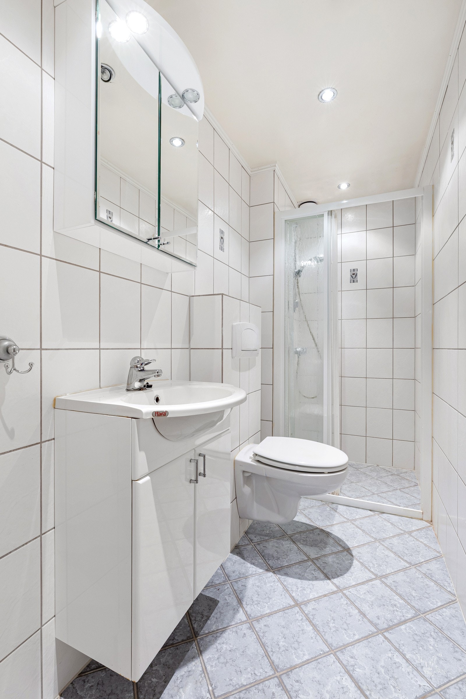 Flislagt gulv med gulvvarme. Flislagte vegger. Himling i lysmalt slett flate med downlights. Gulvstående servantinnredning med profilierte lyse frontr. Heldekkende servant og vegghengt toalett. Innebygget badekar med ett-greps armatur med hånddusj. Galleribilde