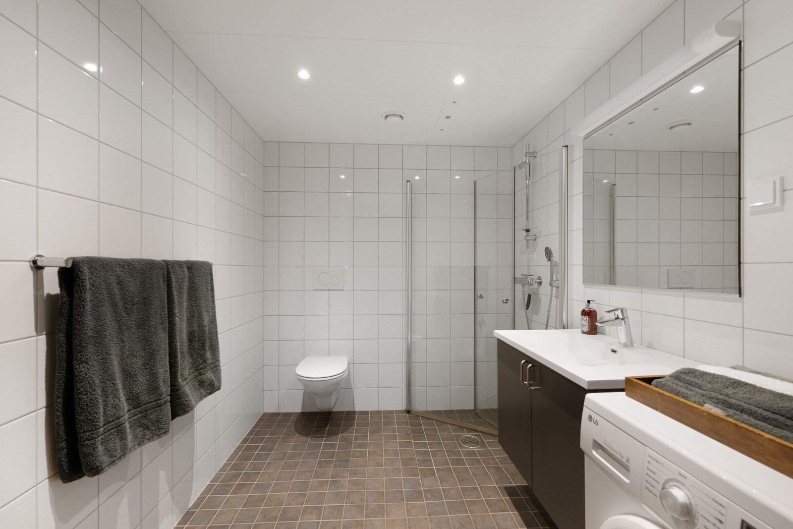 Delikat bad/vaskerom med flislagt gulv med gulvvarme, fliser på vegger og metallplater i himling med downlights. Galleribilde