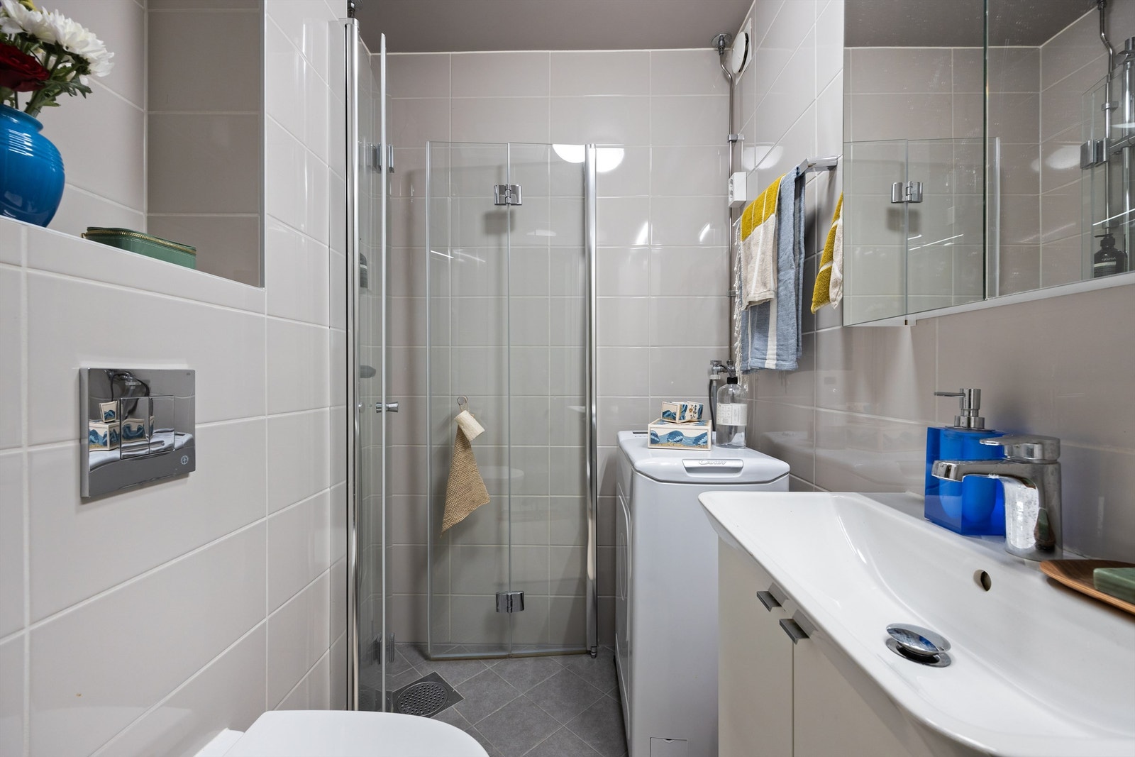 Badet er pent flislagt på gulv og vegger med gulvvarme. Av baderomsinnredning er det vegghengt servantskap med ovenpåliggende servant, speilskap med belysning, vegghengt toalett og dusjhjørne med innfellbare glassdører. Galleribilde