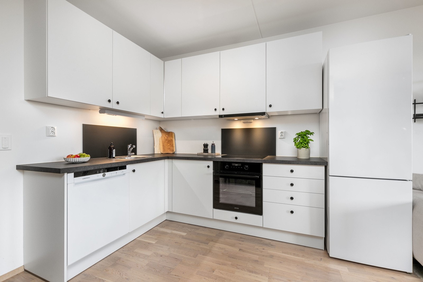 Stilrent og moderne kjøkken fra 2020 med hvite, slette fronter og mørk laminert benkeplate. Kjøkkenet har nedfelt oppvaskkum i rustfritt stål, stilren plate bak koke- og vaskesonen, samt belysning under overskap. Galleribilde