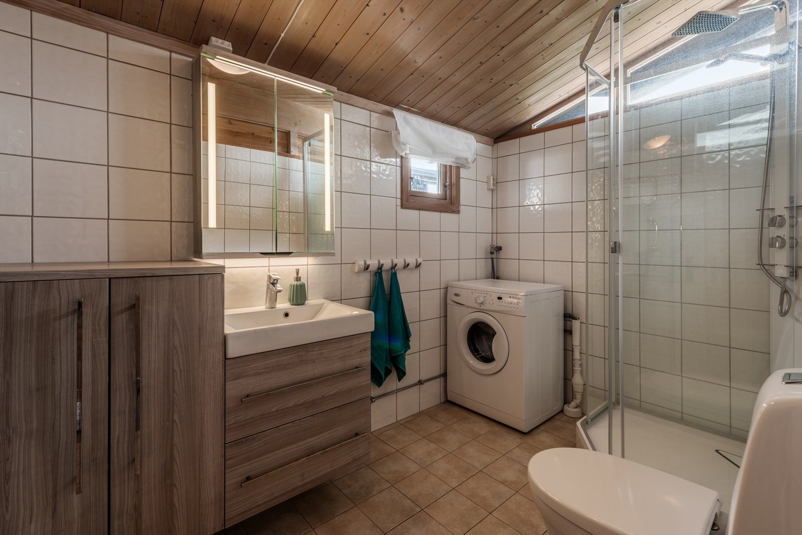 Romslig bad med dusjkabinett og vaskemaskin. Flislagte vegger og gulv gir et stilrent uttrykk. Galleribilde