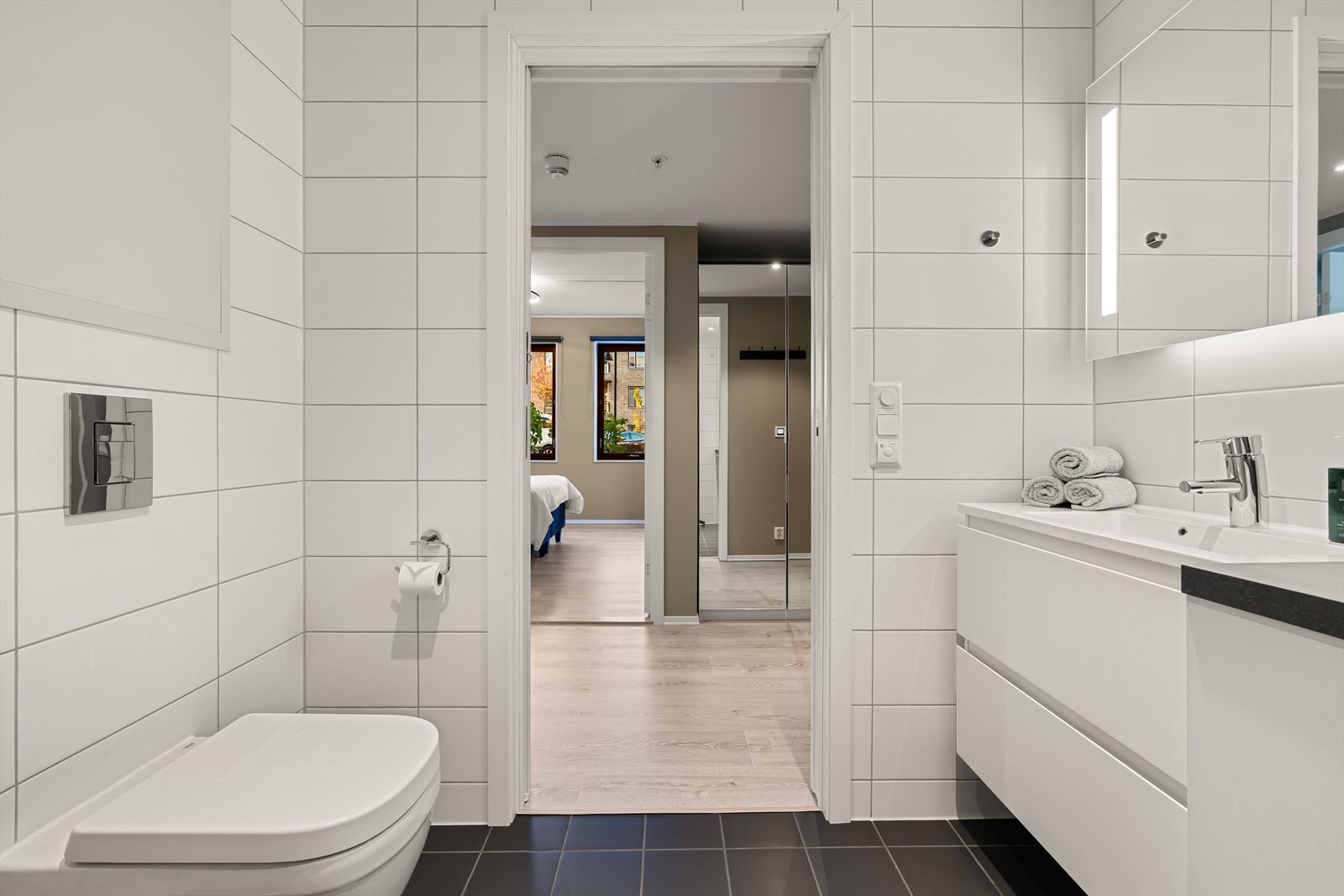 Badet er innredet med vegghengt toalett, dusjhjørne med glassdører, heldekkende servant og speil med integrert belysning. Galleribilde