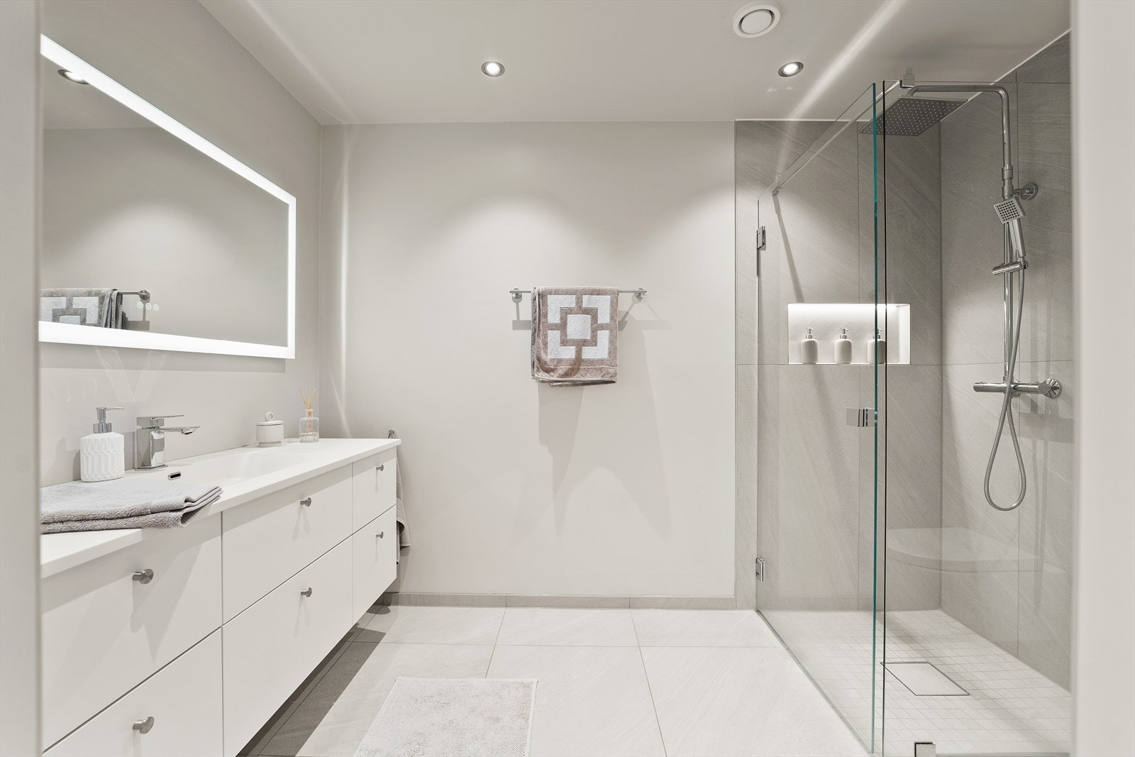 Delikat baderom med fliser på gulv og vegger, vegghengt toalett, romslig HTH-servant og dusjhjørne med praktisk nisje. Galleribilde