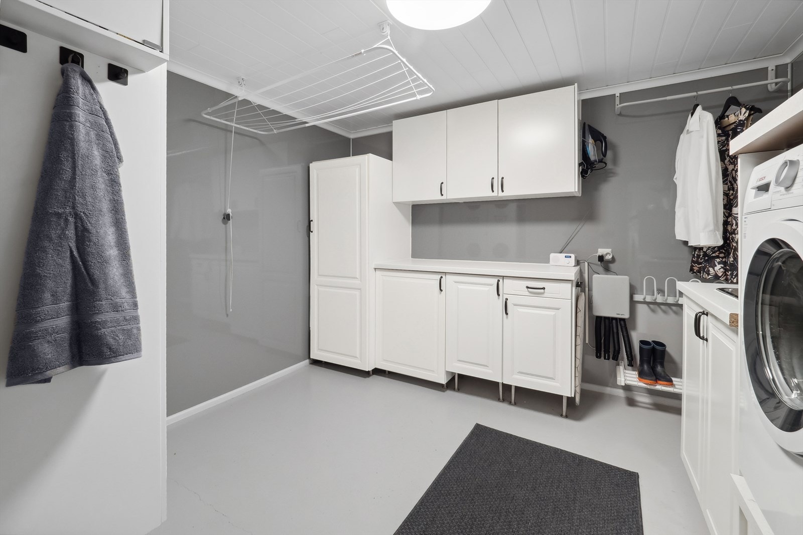 Vaskerom med baderomsplater på veggene og betong på gulv med gulvvarme. Vaskerommet har servant med underskap og opplegg for vaskemaskin. Galleribilde