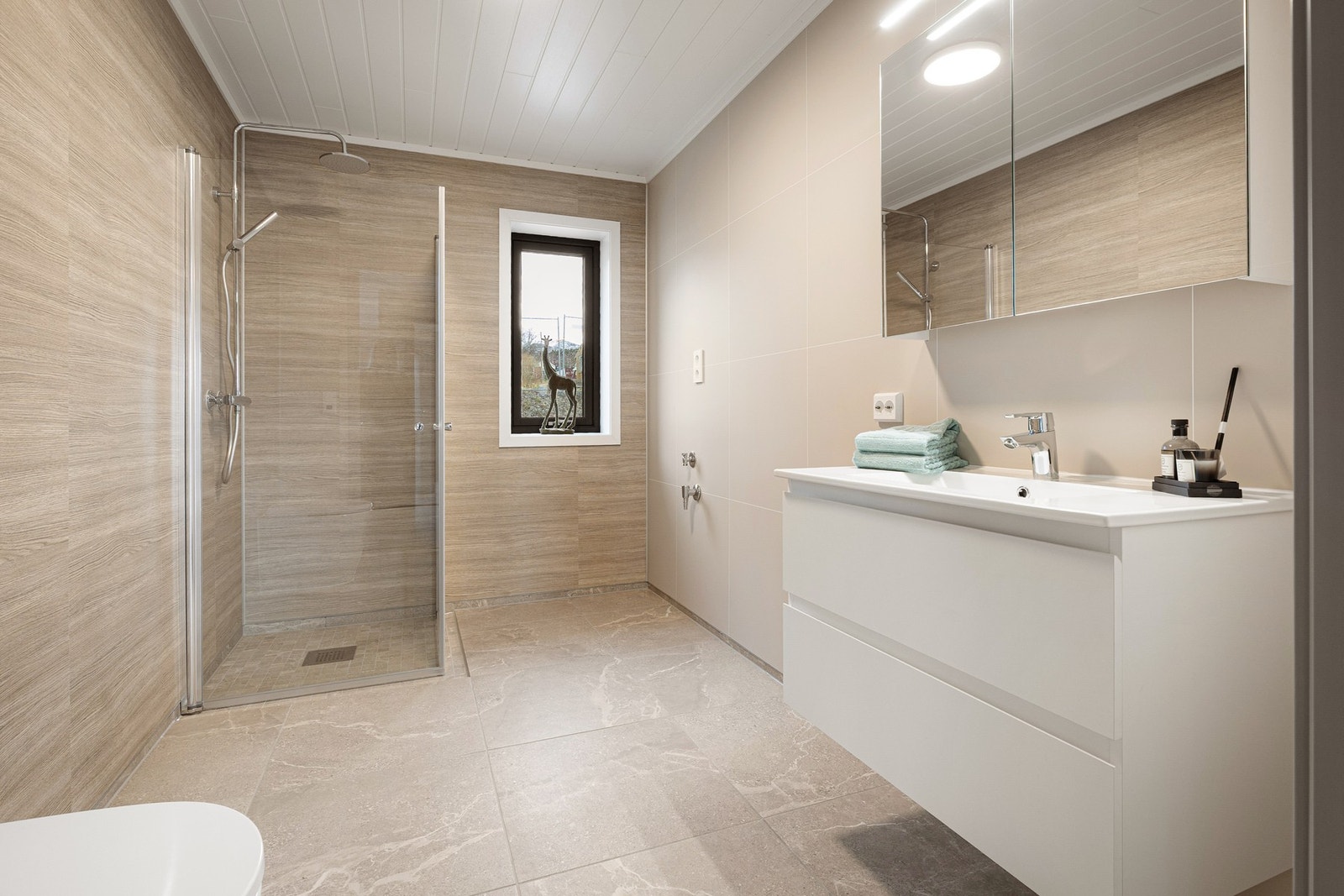 Bad med fliser på gulv og våtromsplater på vegg. Tidløs servantseksjon, vegghengt WC og dusjnisje med glassdører. Galleribilde